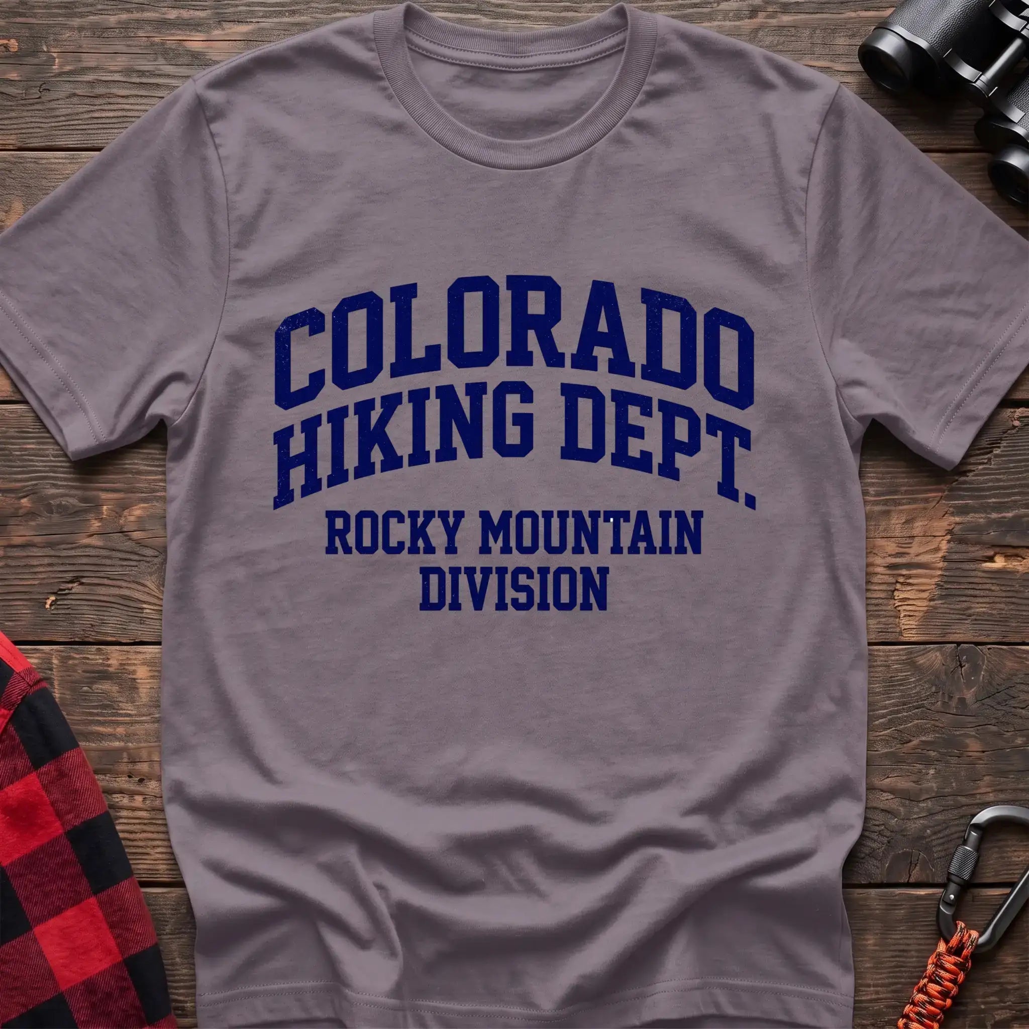 Paragon t-shirt with distressed dark blue collegiate-style text reading “COLORADO HIKING DEPT. ROCKY MOUNTAIN DIVISION,” laid flat on a wooden surface.
