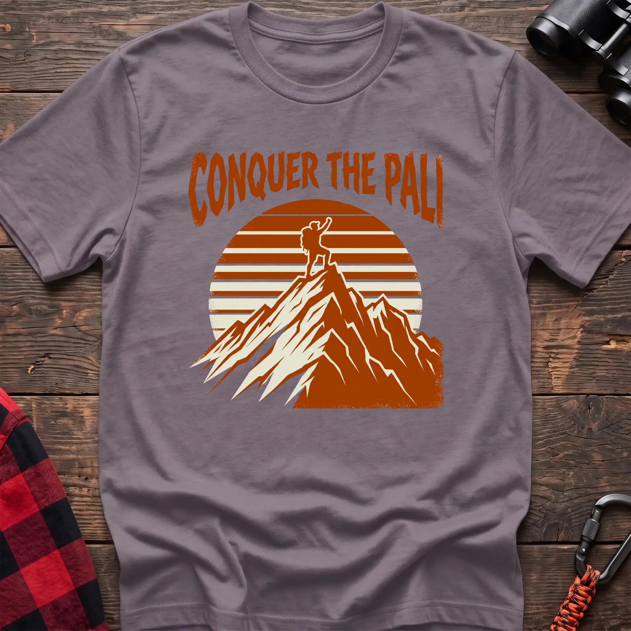 Paragon t-shirt with orange text reading Conquer the Pali above a hiker standing on a mountain peak with a retro sun graphic