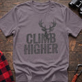 Paragon t-shirt with a distressed dark graphic of a deer’s head and antlers rising from the bold text “CLIMB HIGHER,” laid flat on a wooden surface.
