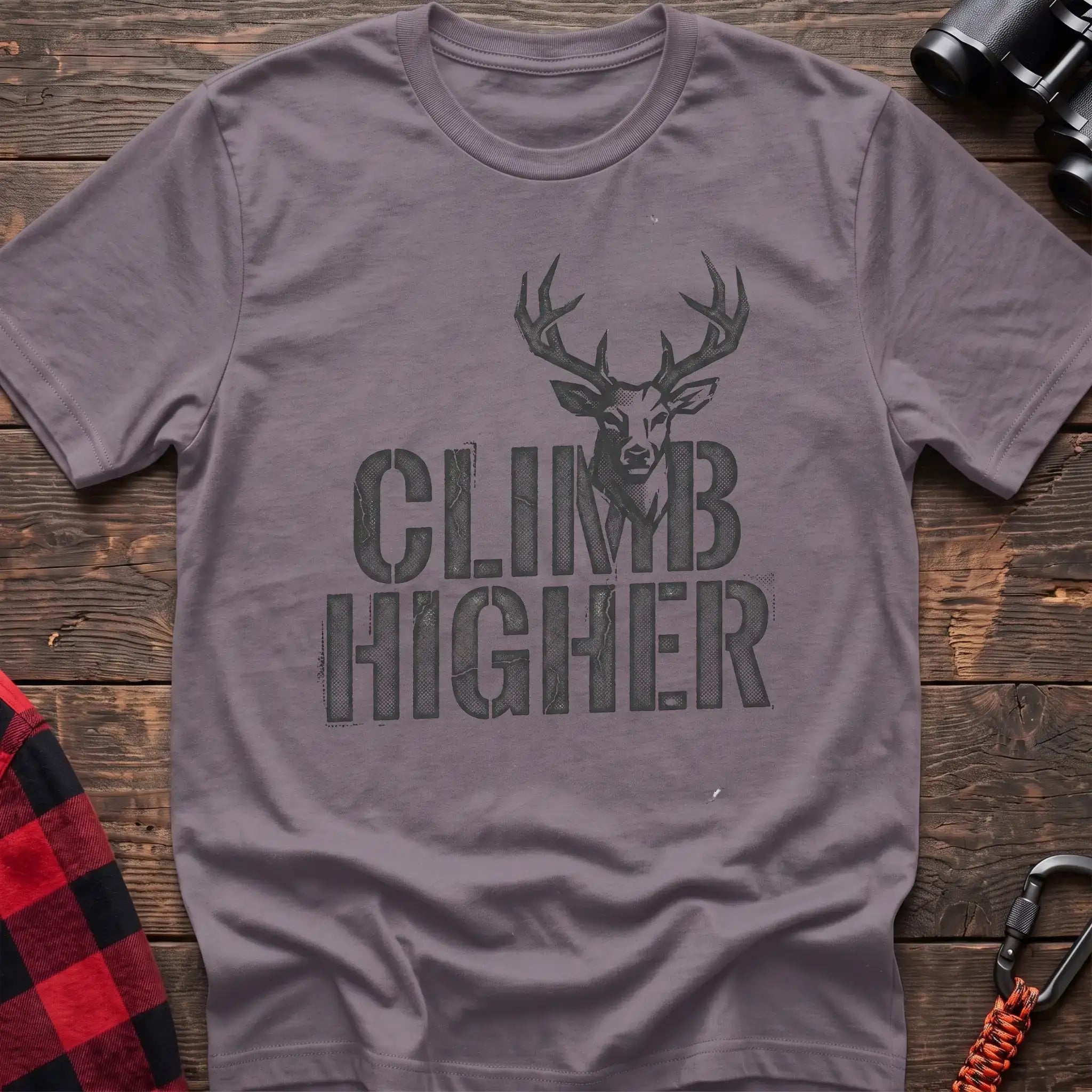 Paragon t-shirt with a distressed dark graphic of a deer’s head and antlers rising from the bold text “CLIMB HIGHER,” laid flat on a wooden surface.