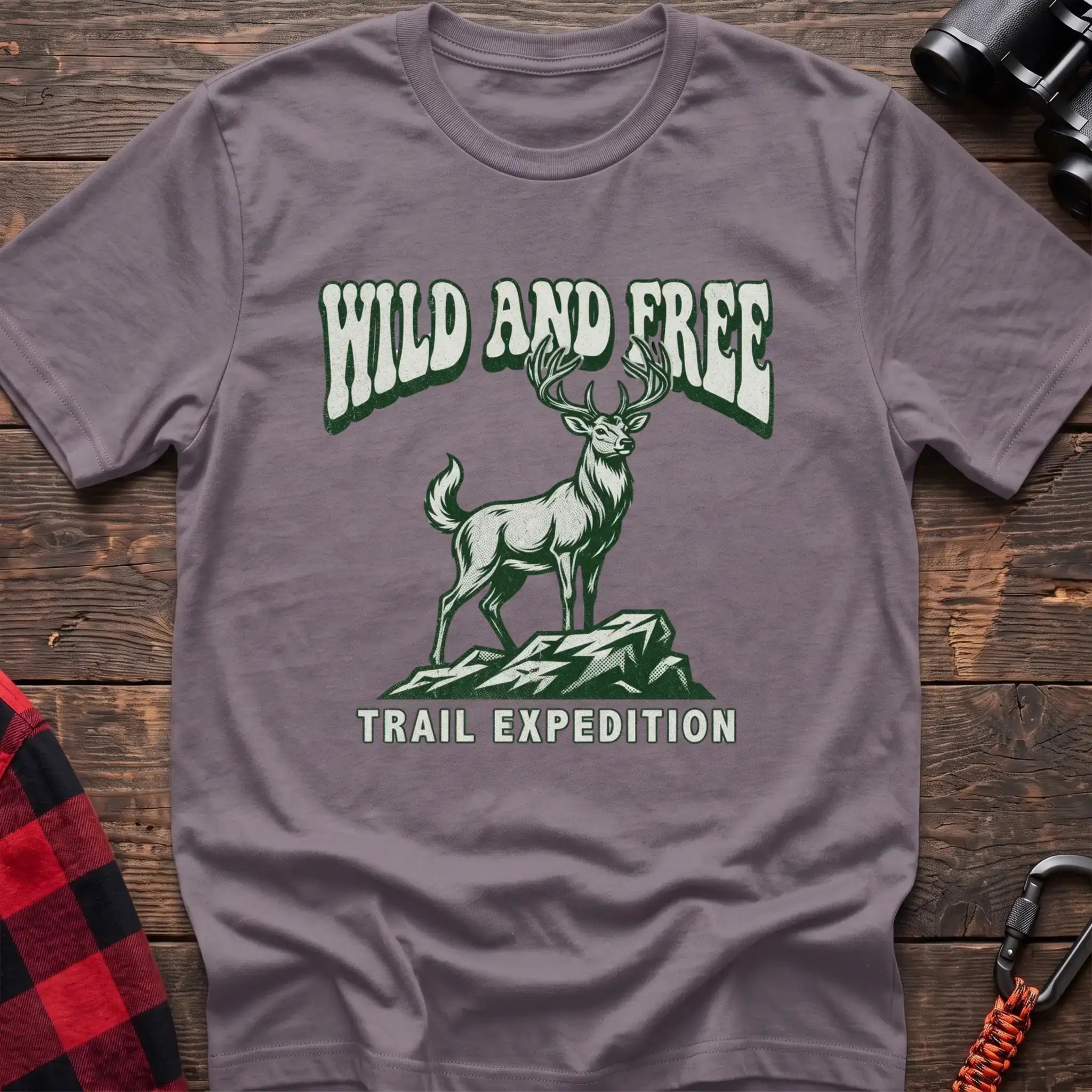Paragon t-shirt with a rustic graphic of a deer standing on a rocky outcrop, with the arched text “WILD AND FREE” above and “TRAIL EXPEDITION” below, laid flat on a wooden surface.