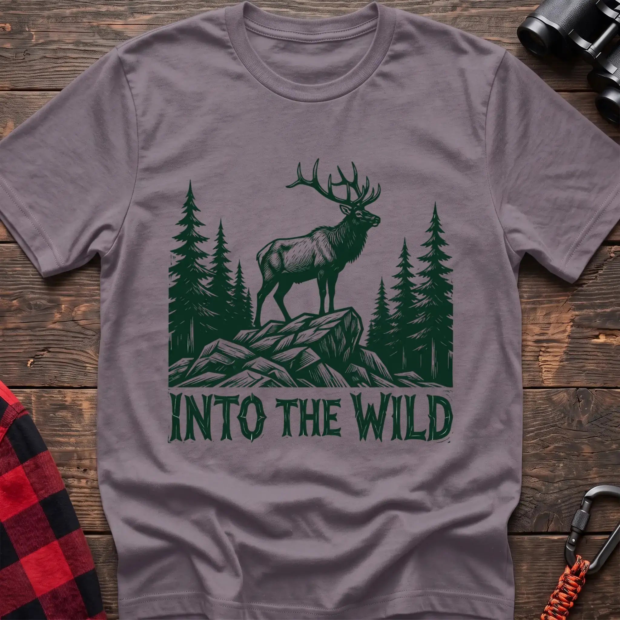 Paragon t-shirt with a dark green illustration of an elk standing on a rocky outcrop among pine trees, with the text “INTO THE WILD” printed below, laid flat on a wooden surface.