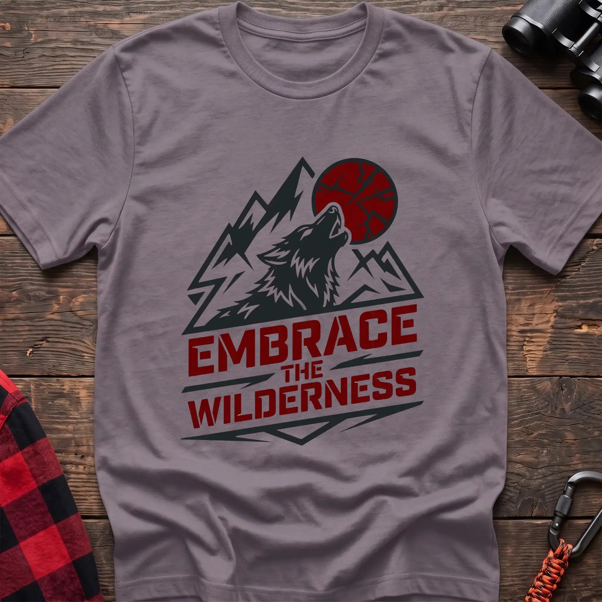 Paragon t-shirt with a dark graphic of a howling wolf against mountain peaks and a red sun, with the bold red text “EMBRACE THE WILDERNESS” below, laid flat on a wooden surface.