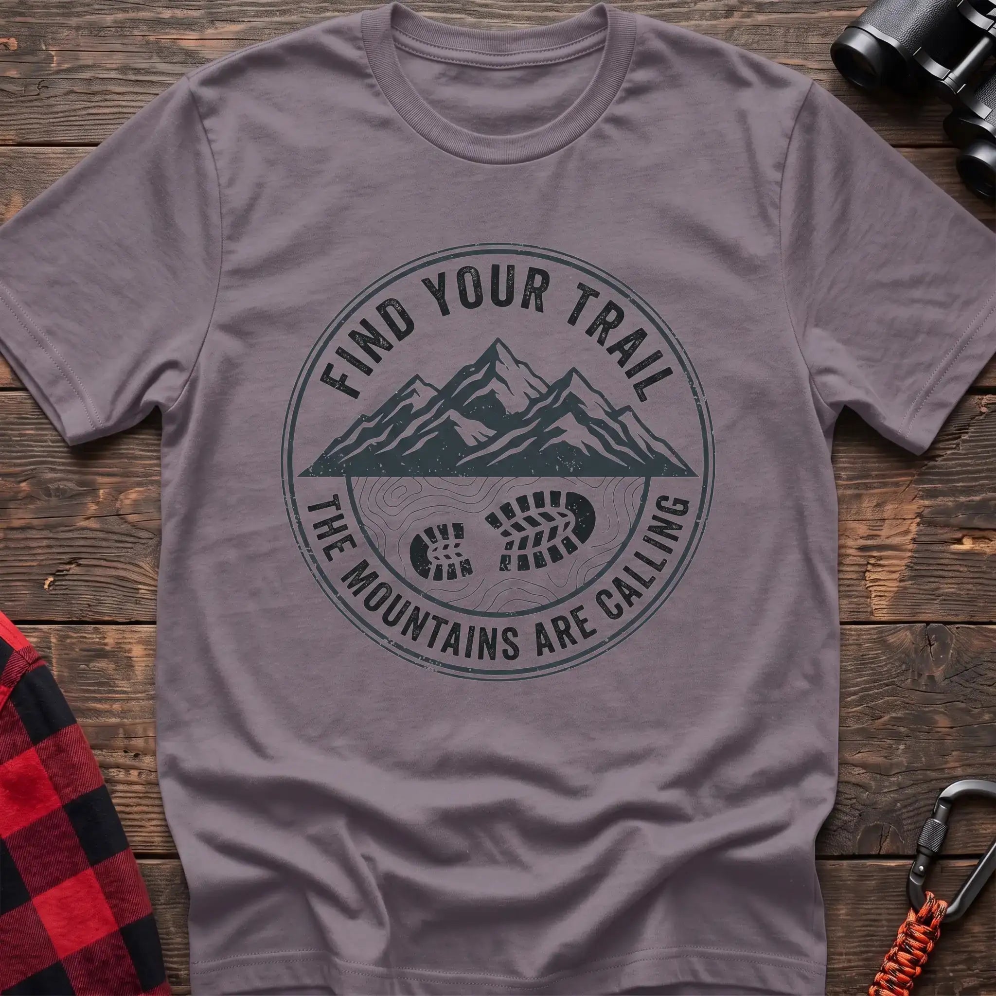 Paragon t-shirt with a circular dark gray badge design featuring mountain peaks above a hiking boot print on topographic lines, with the text “FIND YOUR TRAIL” and “THE MOUNTAINS ARE CALLING,” laid flat on a wooden surface.