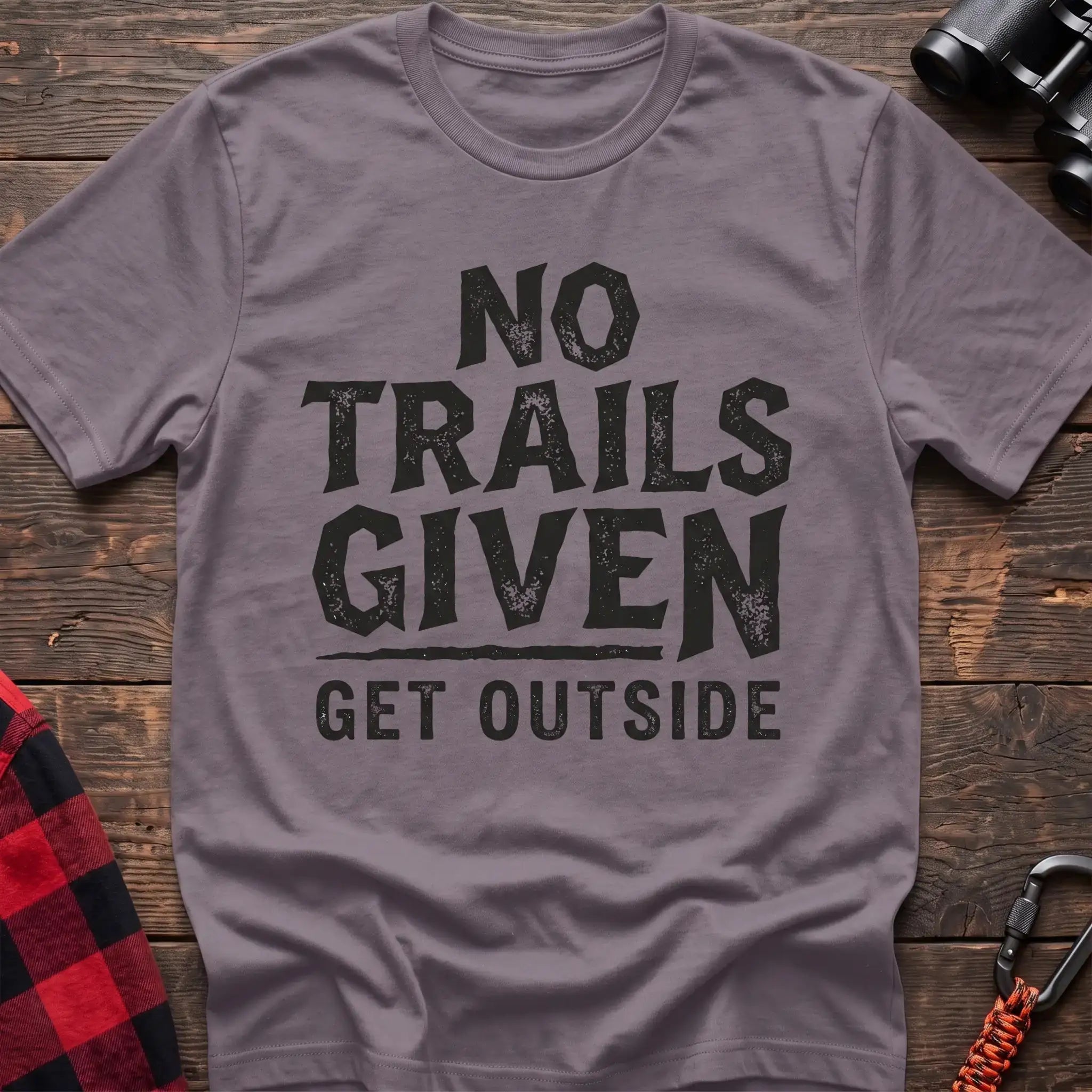 Paragon t-shirt with distressed black text reading “NO TRAILS GIVEN” and “GET OUTSIDE” below it, laid flat on a wooden surface.
