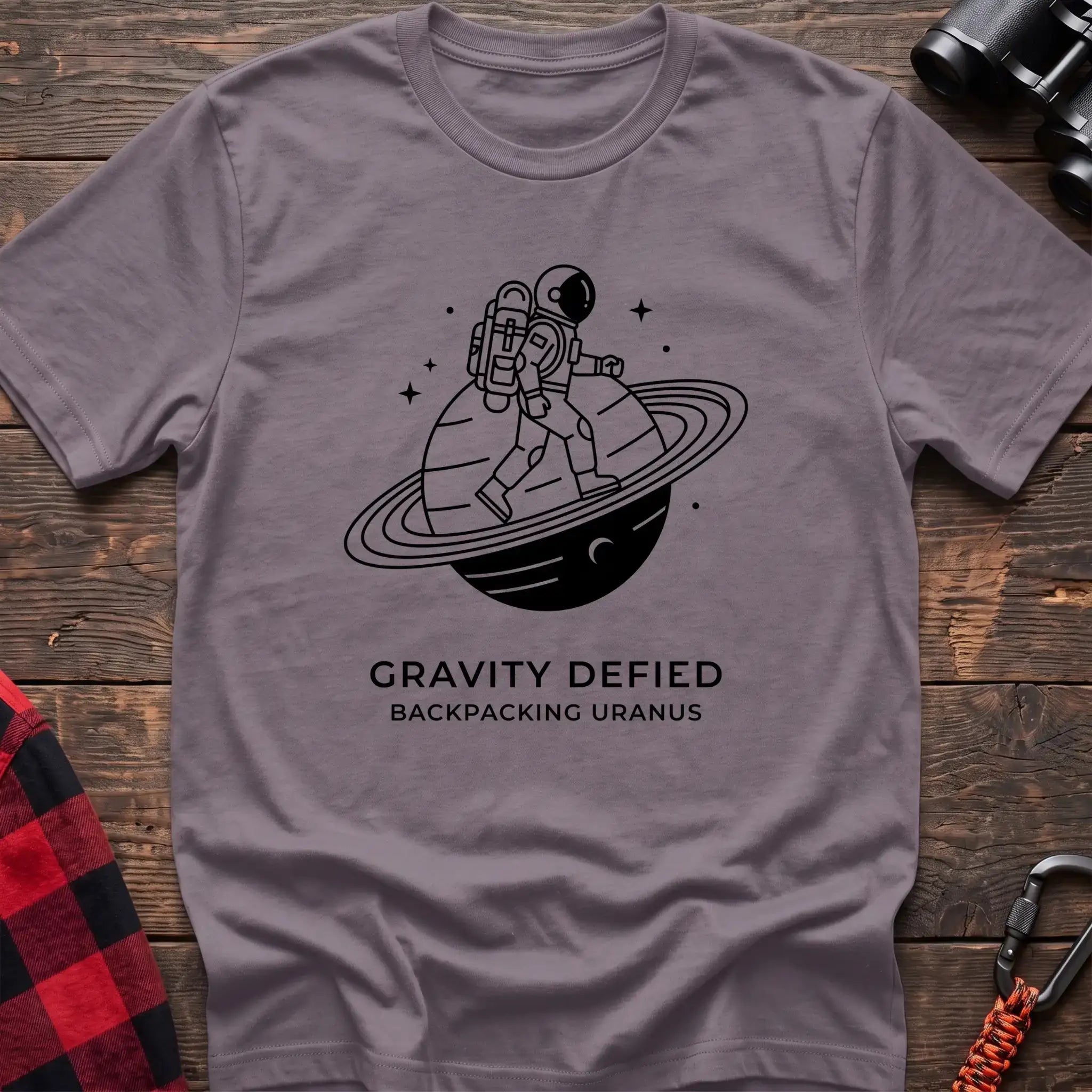 Paragon crew-neck t-shirt with black line-art graphic of an astronaut backpacking across a ringed planet surrounded by stars, with the text “GRAVITY DEFIED / BACKPACKING URANUS” printed below, laid flat on a wooden surface.