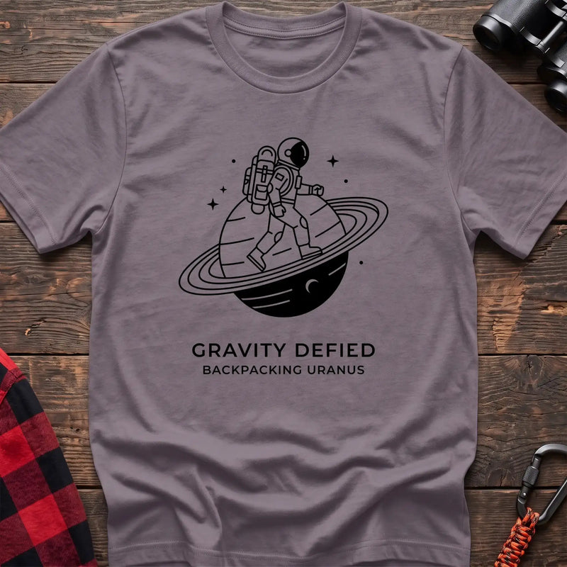 Paragon crew-neck t-shirt with black line-art graphic of an astronaut backpacking across a ringed planet surrounded by stars, with the text “GRAVITY DEFIED / BACKPACKING URANUS” printed below, laid flat on a wooden surface.
