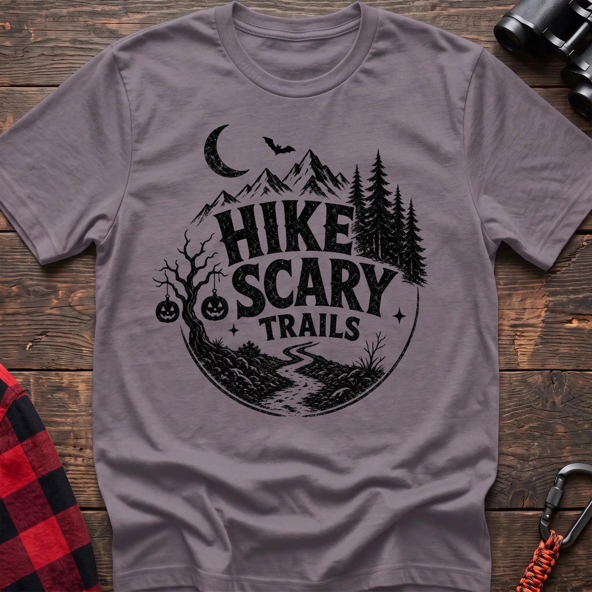 Paragon cotton t-shirt with vintage Hike Scary Trails graphic of mountains, a crescent moon, bat, jack-o-lanterns, and a winding trail, flat lay on rustic wood with hiking gear