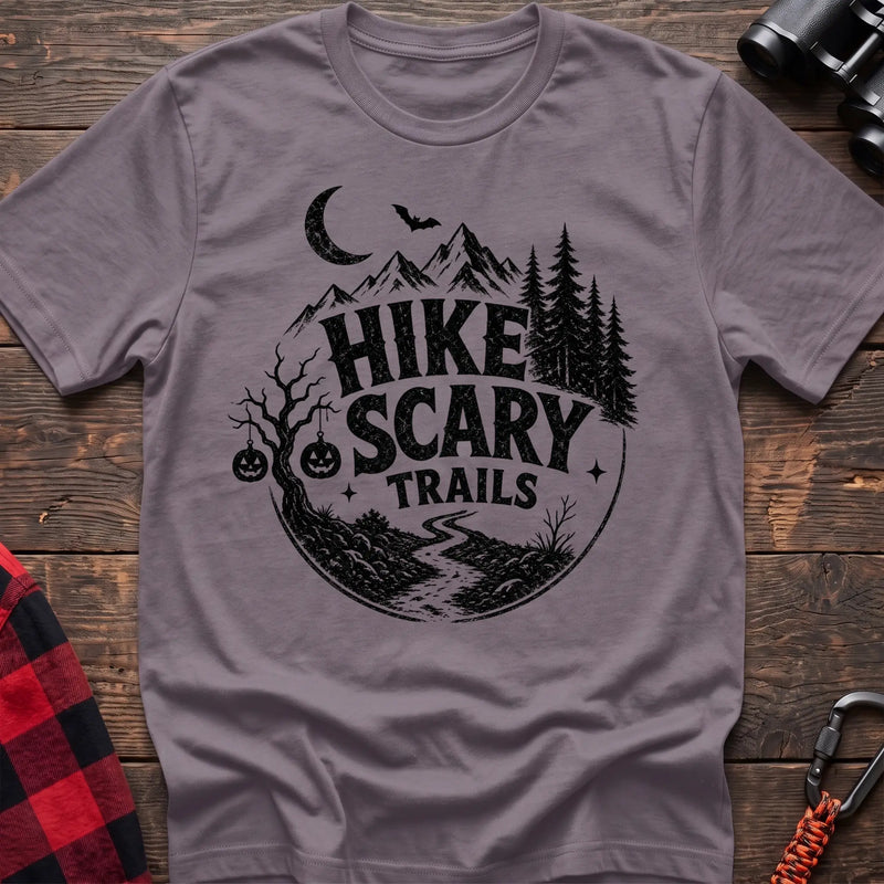 Paragon cotton t-shirt with vintage Hike Scary Trails graphic of mountains, a crescent moon, bat, jack-o-lanterns, and a winding trail, flat lay on rustic wood with hiking gear