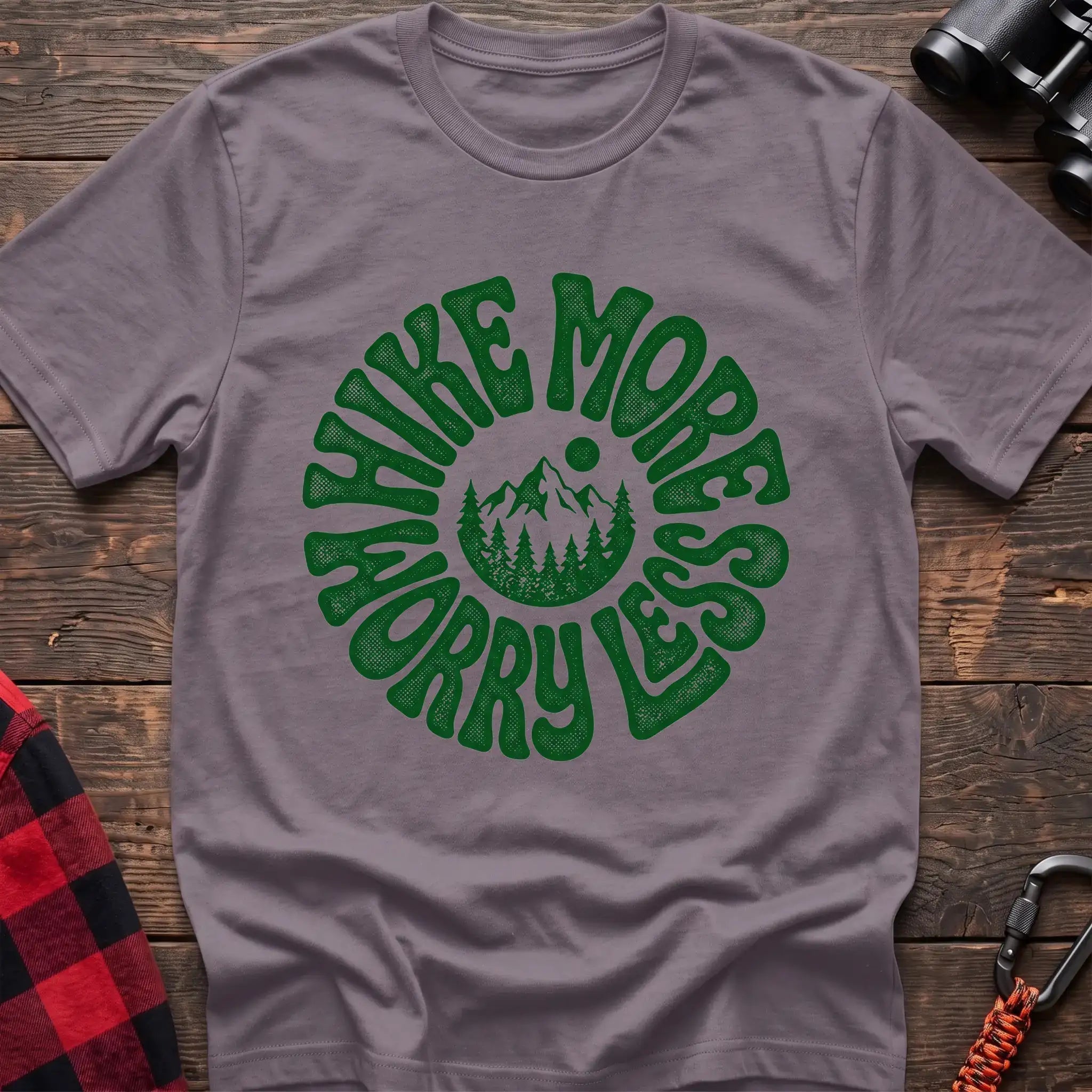 Paragon t-shirt with a circular green design featuring mountains and pine trees in the center, surrounded by the text “HIKE MORE WORRY LESS,” laid flat on a wooden surface.
