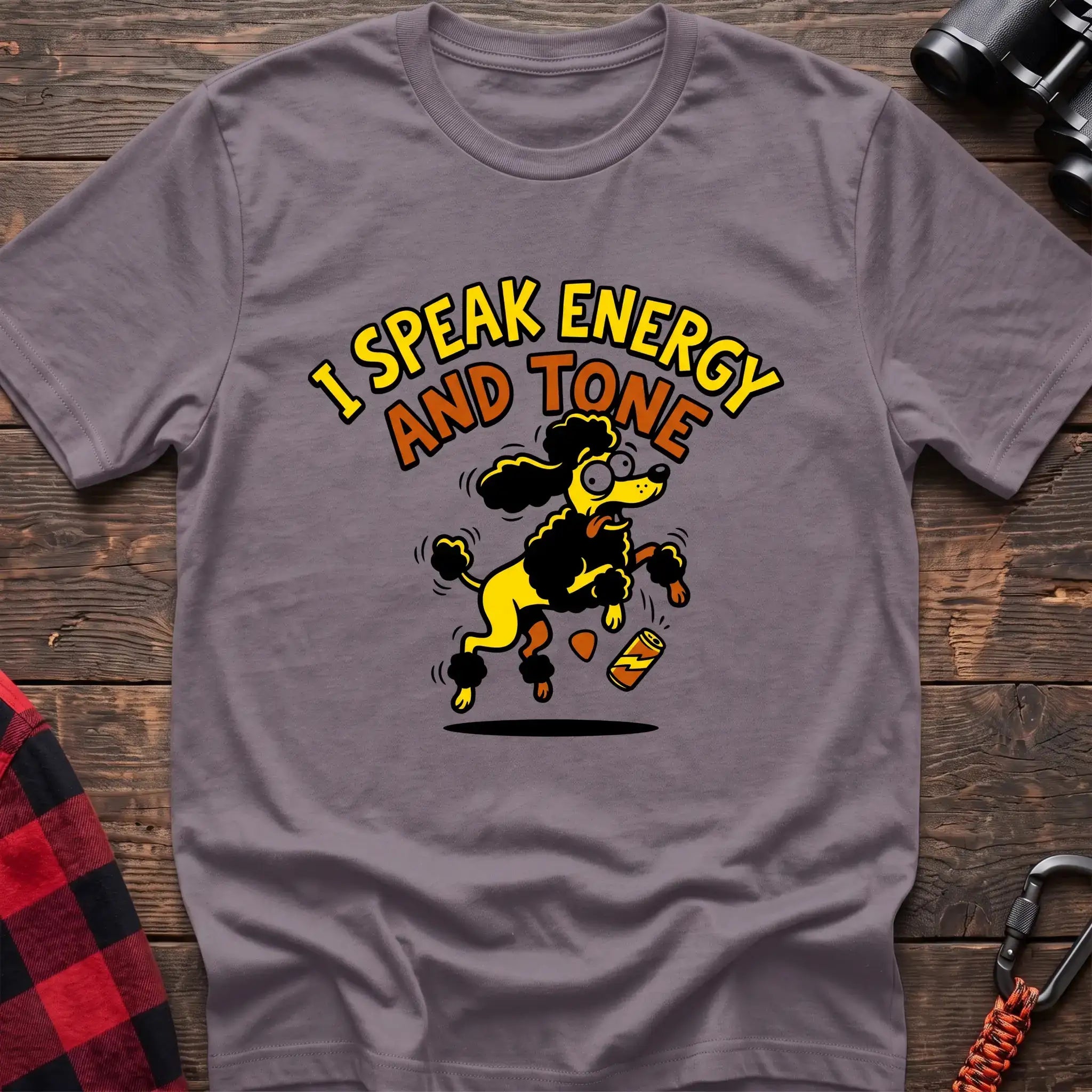 Paragon t-shirt with a cartoon yellow and black poodle jumping excitedly next to a falling energy drink can, with the arched text “I SPEAK ENERGY AND TONE,” laid flat on a wooden surface.