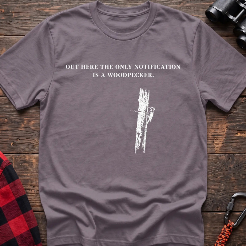 Paragon cotton t-shirt with a woodpecker graphic and Out Here the Only Notification Is a Woodpecker text on rustic wood
