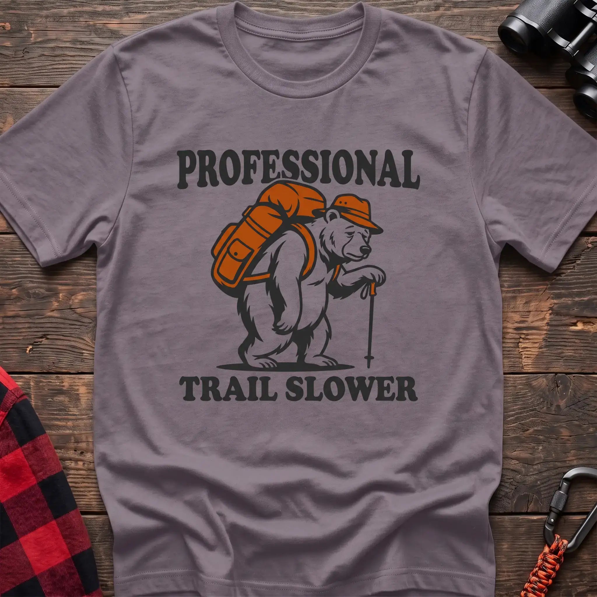 Paragon t-shirt with a cartoon polar bear wearing a backpack and hat while holding a hiking stick, with the text “PROFESSIONAL TRAIL SLOWER,” laid flat on a wooden surface.