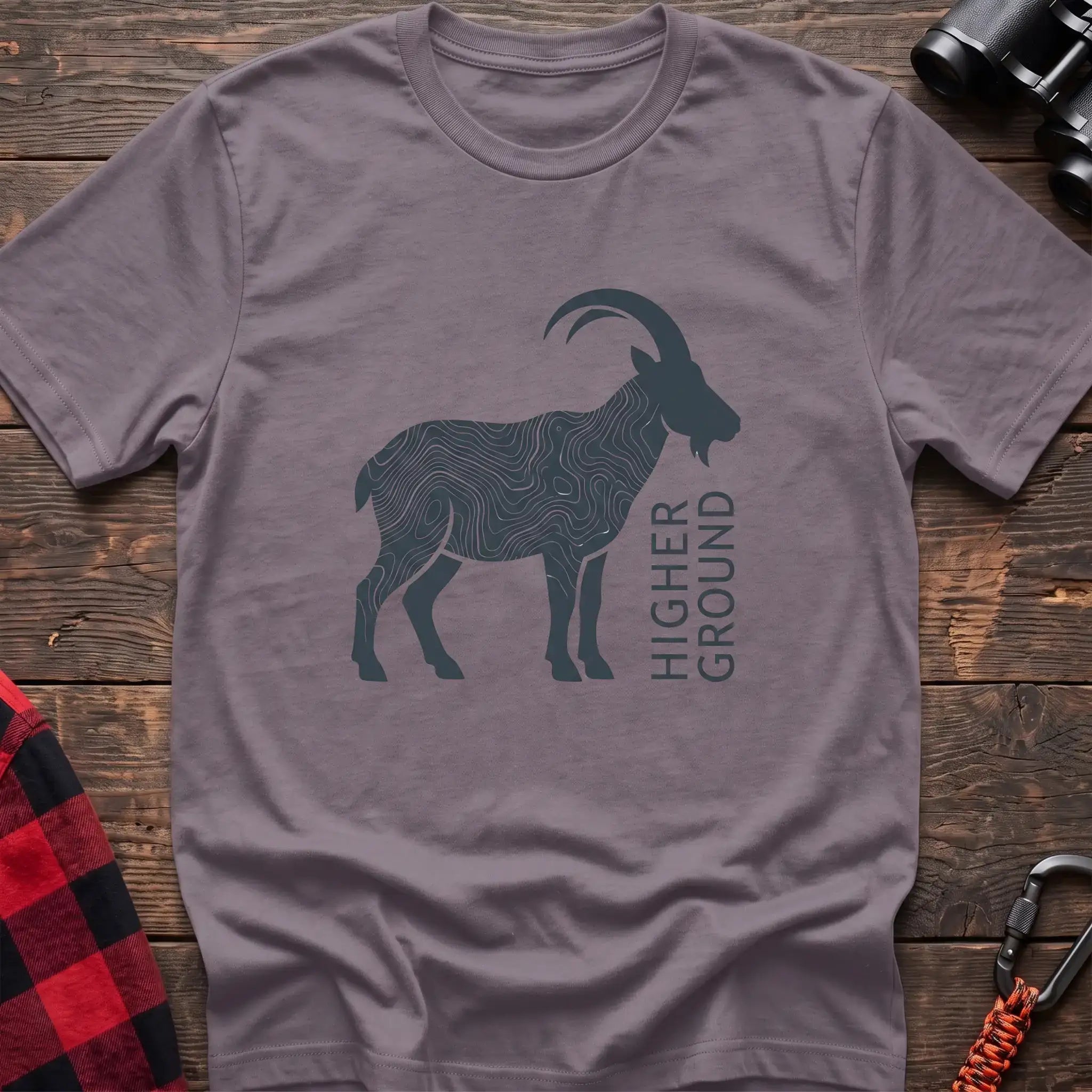 Paragon t-shirt with a dark silhouette of a mountain goat filled with topographic contour lines, and the vertical text “HIGHER GROUND” printed beside it, laid flat on a wooden surface.