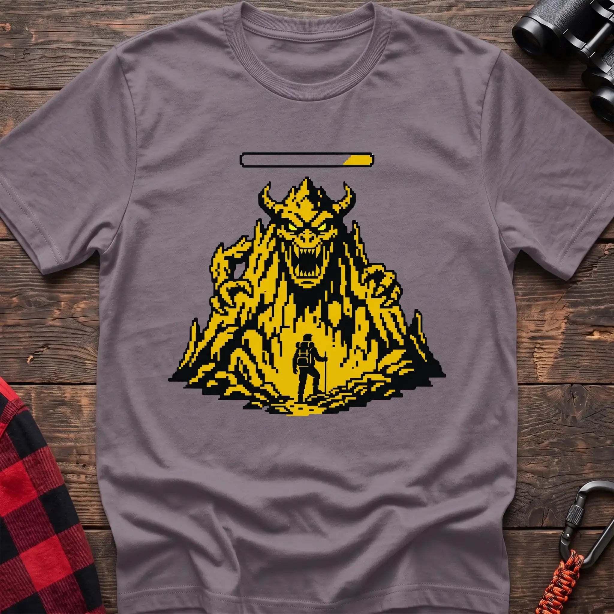 Paragon t-shirt with a pixel-art illustration of a hiker facing a large horned mountain monster with a health bar above it, laid flat on a wooden surface.