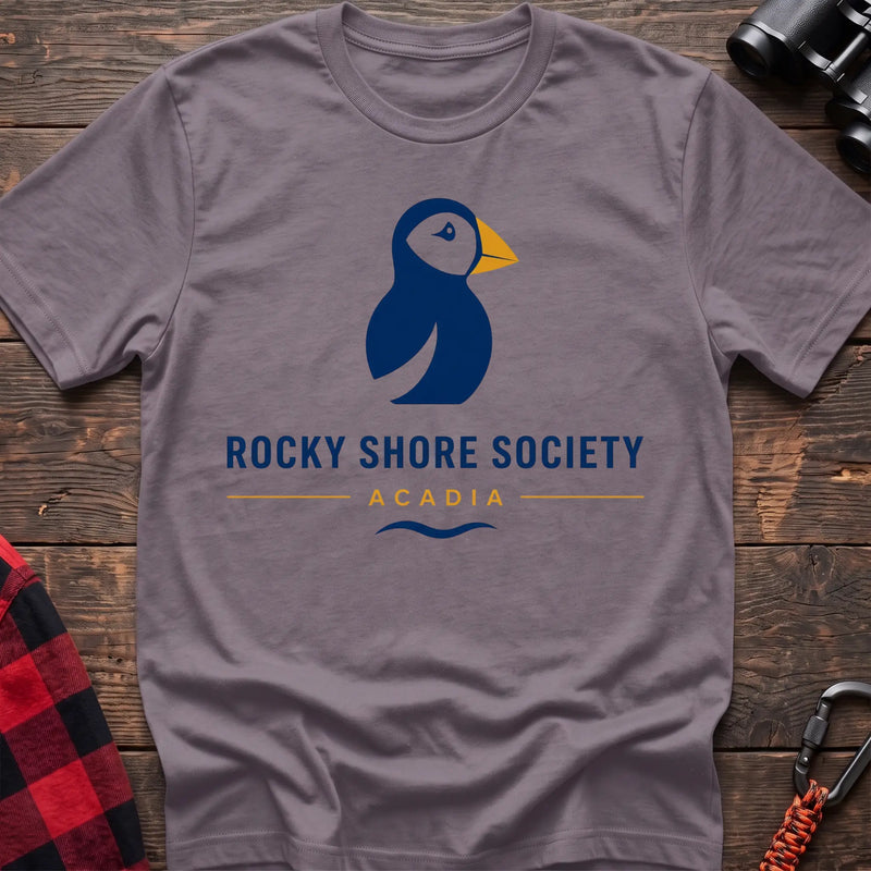 Paragon cotton t-shirt with a navy puffin logo, yellow beak, and Rocky Shore Society Acadia text on a rustic wood background