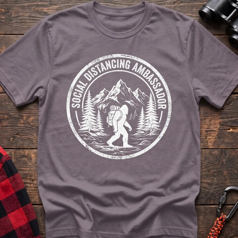 Paragon t-shirt with white circular badge graphic of Bigfoot hiking with a backpack through mountains and the text Social Distancing Ambassador