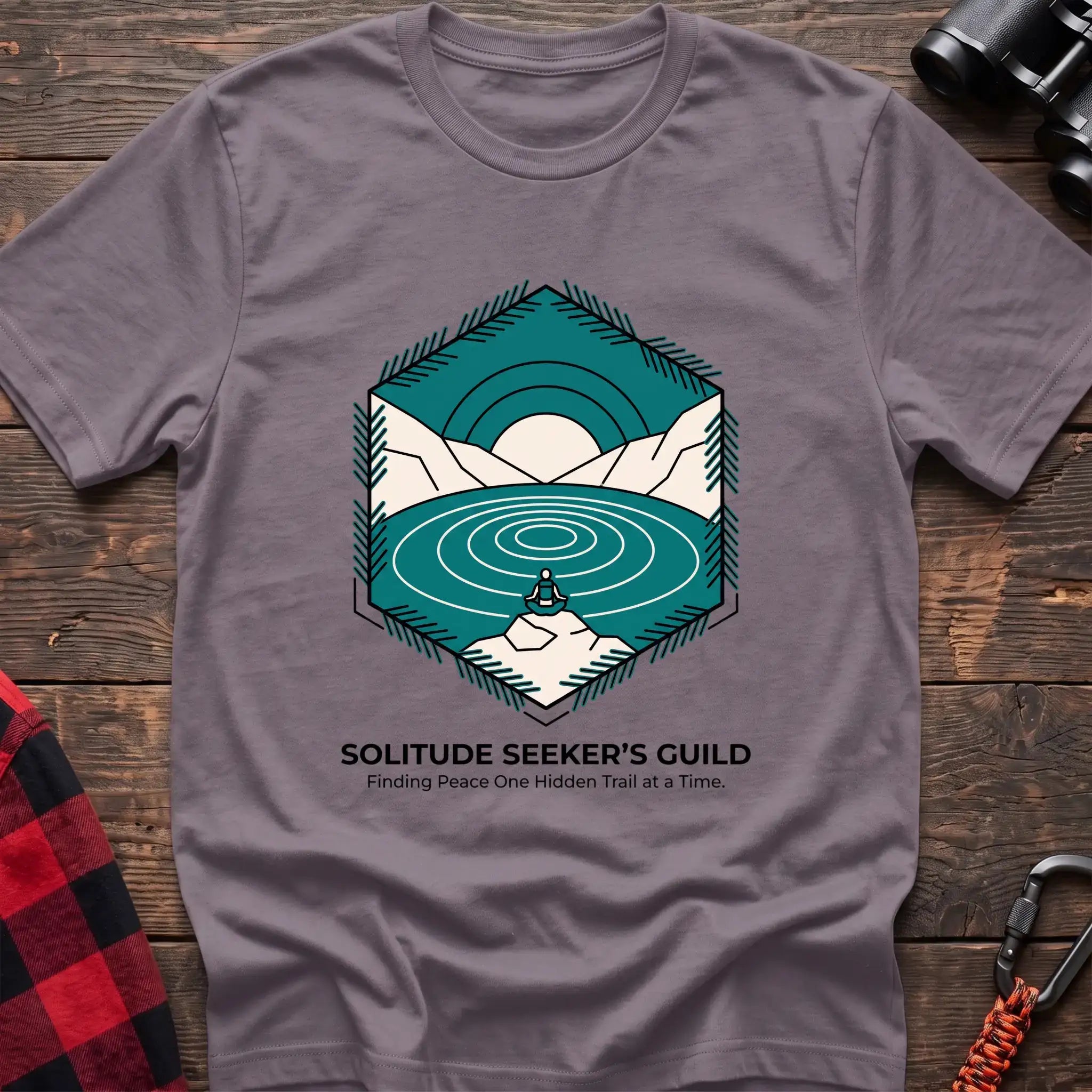 Paragon t-shirt with a hexagonal illustration of a person meditating by a mountain lake, with the text “SOLITUDE SEEKER’S GUILD” and “Finding Peace One Hidden Trail at a Time,” laid flat on a wooden surface.