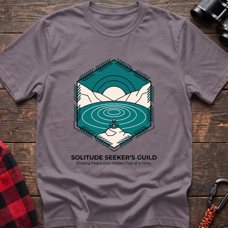 Paragon t-shirt with a hexagonal illustration of a person meditating by a mountain lake, with the text “SOLITUDE SEEKER’S GUILD” and “Finding Peace One Hidden Trail at a Time,” laid flat on a wooden surface.