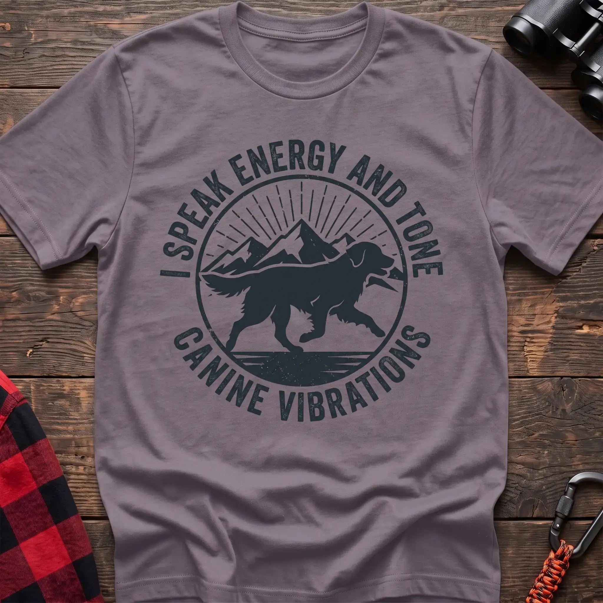 Paragon t-shirt with a circular dark gray badge design featuring a running dog carrying a stick against mountain peaks and sun rays, with the text “I SPEAK ENERGY AND TONE” and “CANINE VIBRATIONS,” laid flat on a wooden surface.