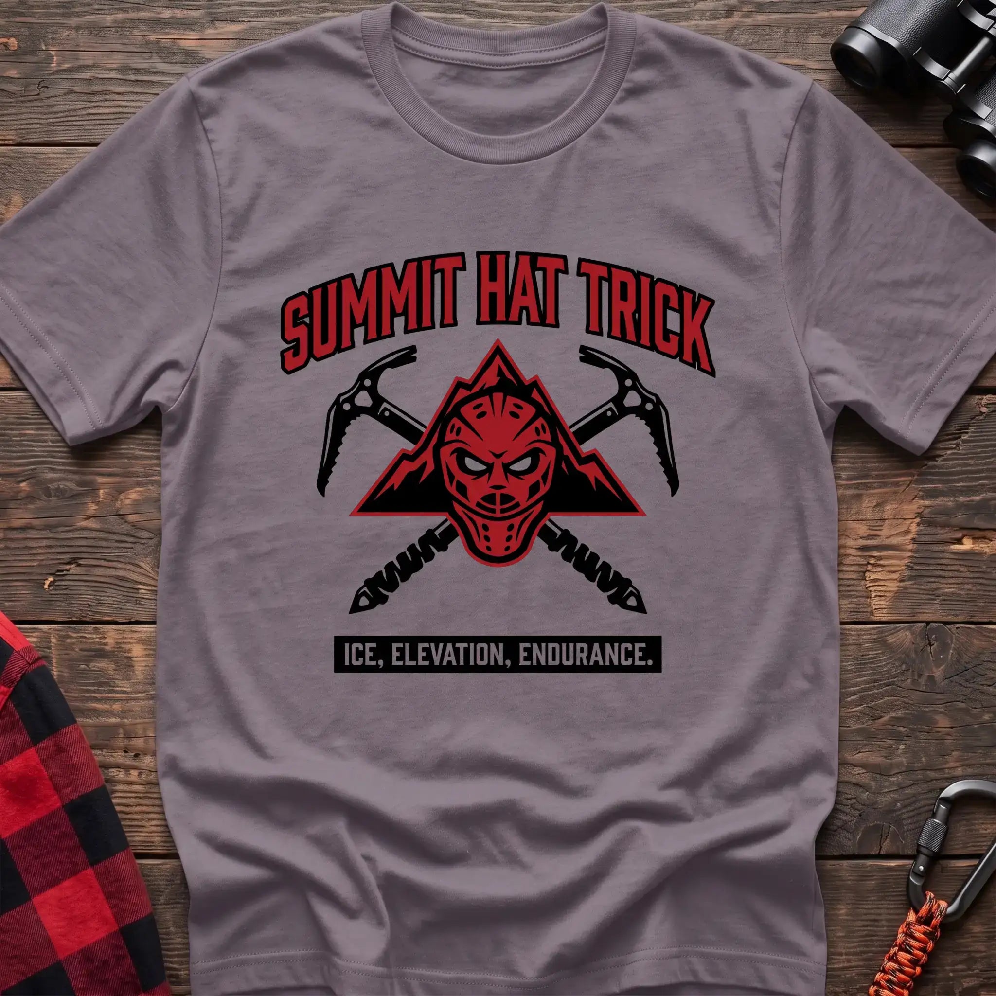 Paragon t-shirt with a red mountain-themed goalie mask and crossed ice axes, with the text “SUMMIT HAT TRICK” and “ICE, ELEVATION, ENDURANCE,” laid flat on a wooden surface.