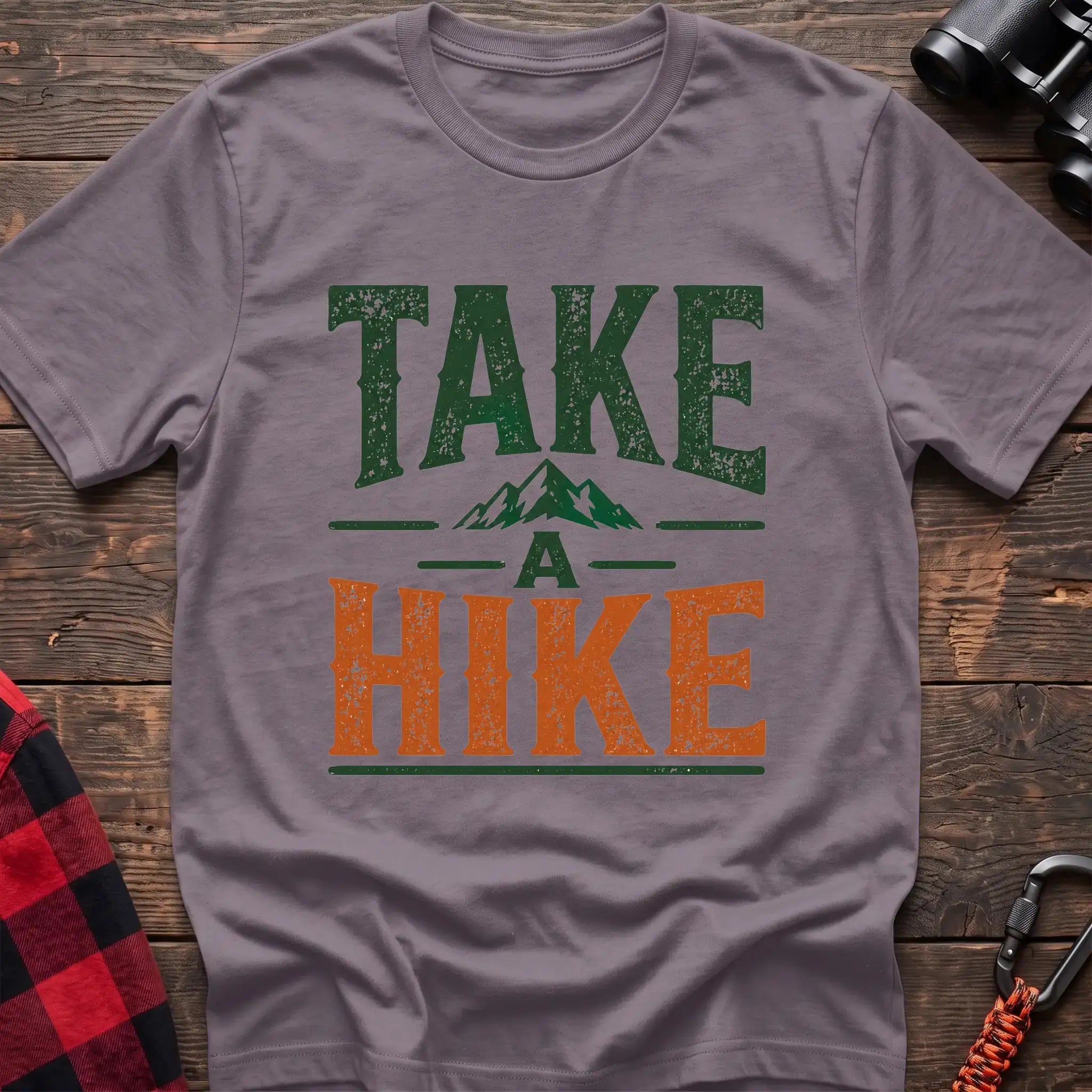 Paragon t-shirt with distressed green and orange text reading “TAKE A HIKE” with a small mountain graphic between the words, laid flat on a wooden surface.