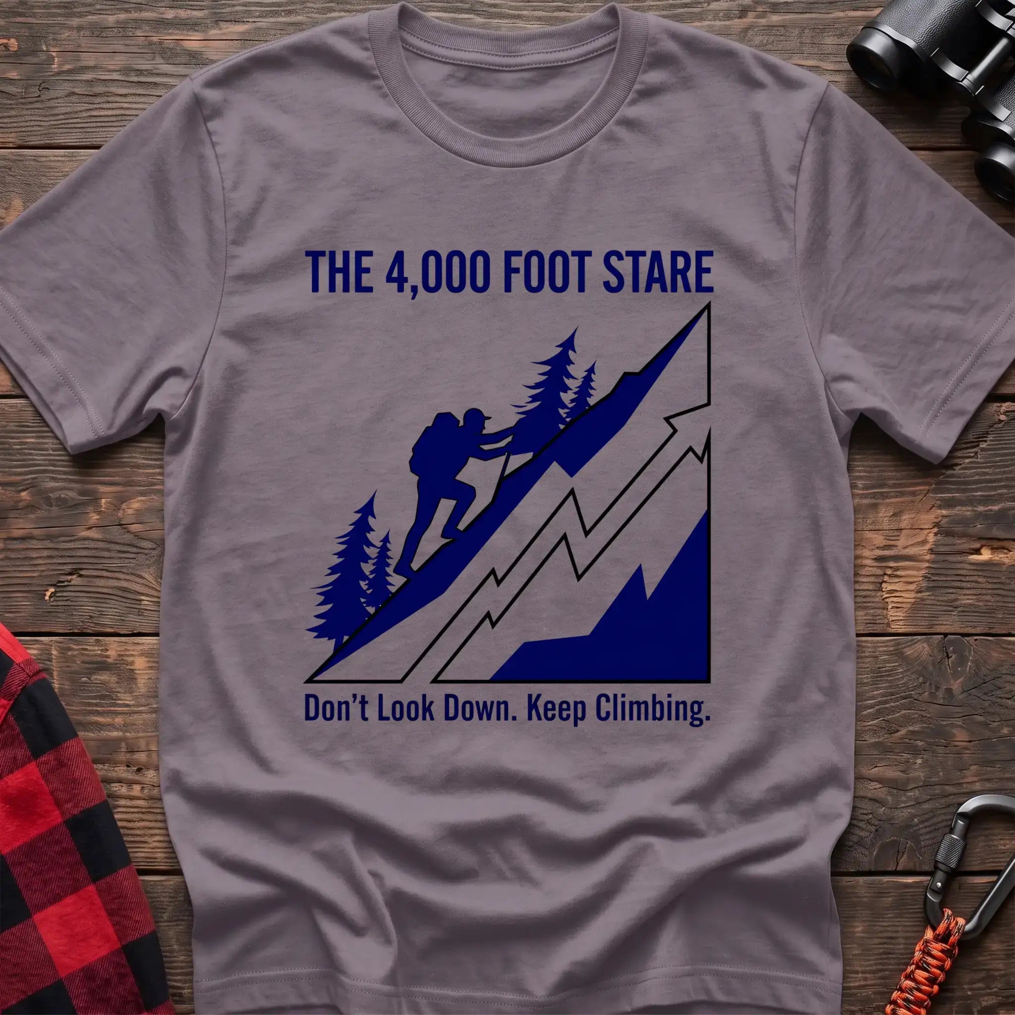 Paragon t-shirt with a blue illustration of a hiker climbing a steep mountain, with the text “THE 4,000 FOOT STARE” and “Don’t Look Down. Keep Climbing,” laid flat on a wooden surface.