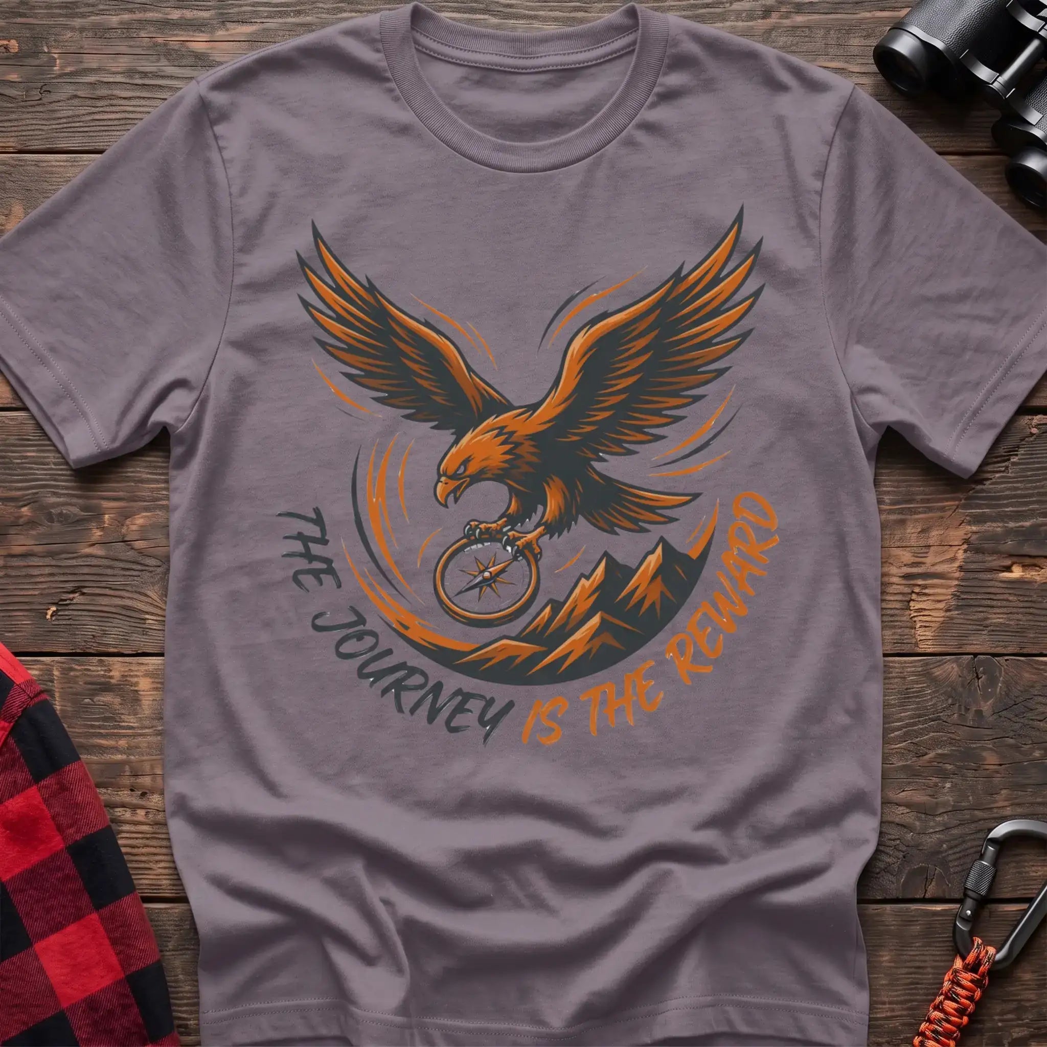 Paragon t-shirt with an orange and dark graphic of a soaring eagle holding a compass above mountains, with the curved text “THE JOURNEY IS THE REWARD,” laid flat on a wooden surface.