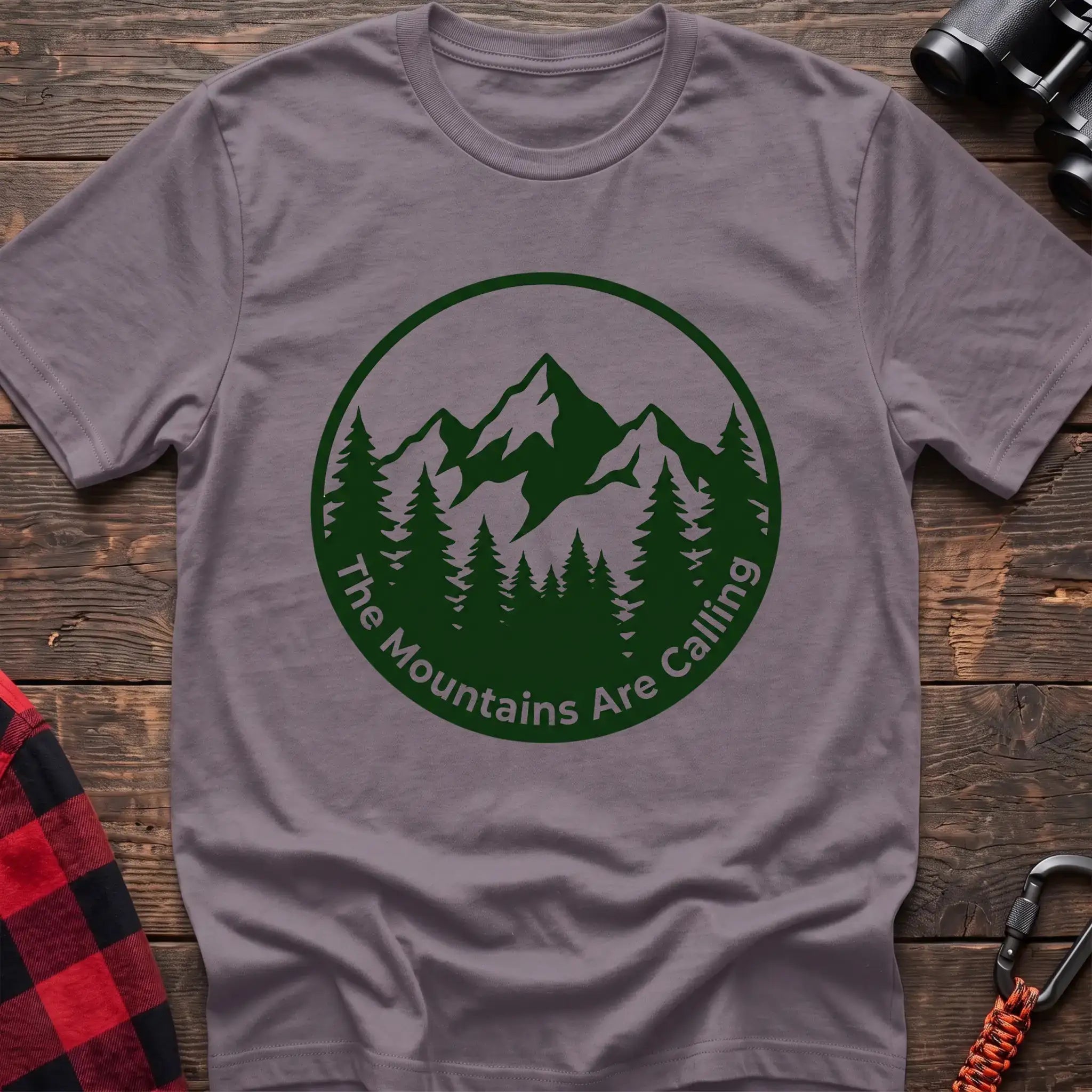 Paragon t-shirt with a circular dark green design featuring mountain peaks and pine trees, with the text “The Mountains Are Calling,” laid flat on a wooden surface.