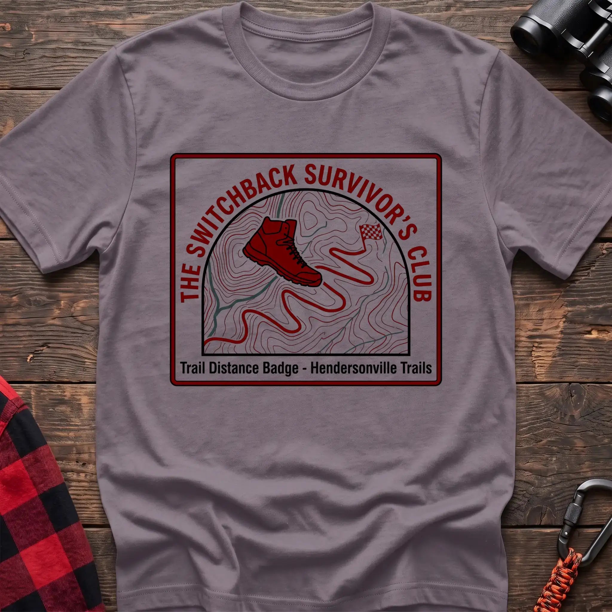 Paragon t-shirt with a square badge design featuring a red hiking boot on a topographic map with a winding trail, and the text “THE SWITCHBACK SURVIVOR’S CLUB” and “Trail Distance Badge - Hendersonville Trails,” laid flat on a wooden surface.