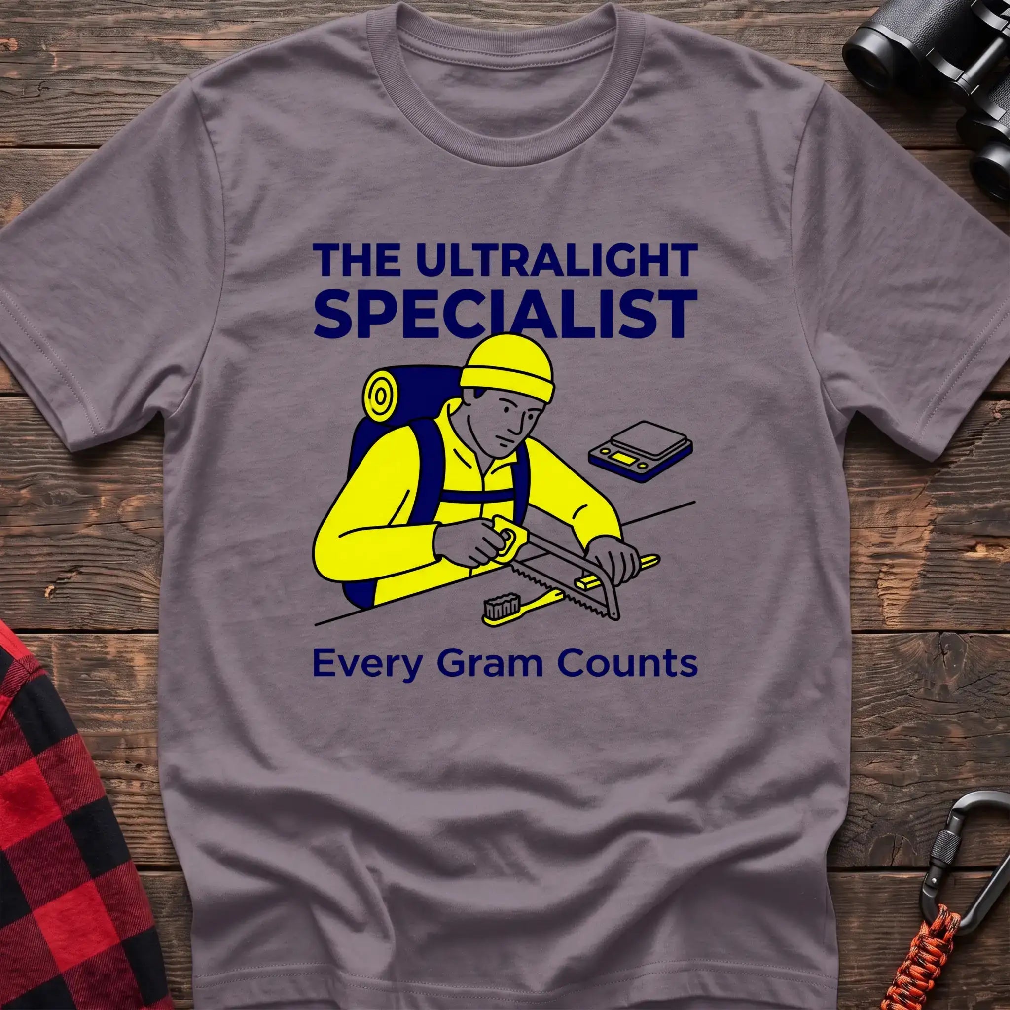Paragon t-shirt with an illustration of a hiker sawing a toothbrush on a table next to a scale, with the text “THE ULTRALIGHT SPECIALIST” and “Every Gram Counts,” laid flat on a wooden surface.