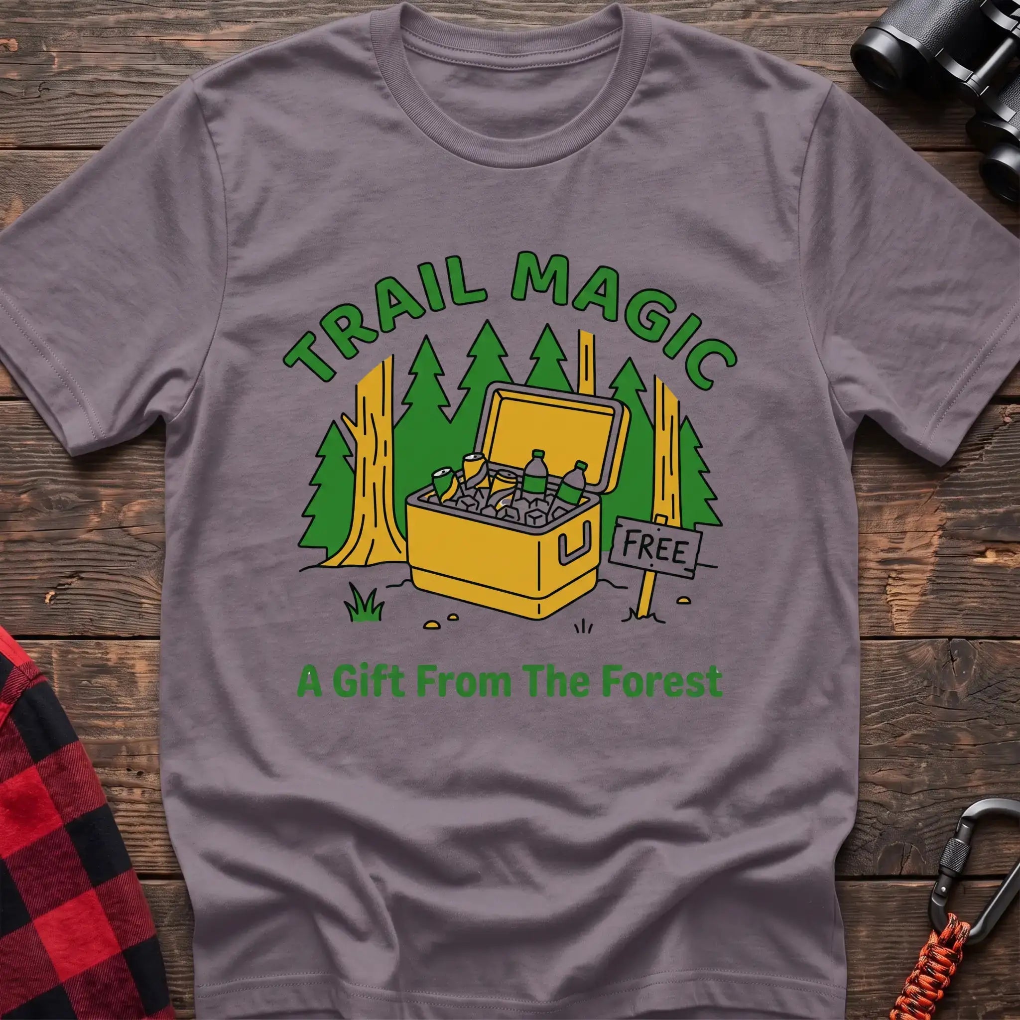 Paragon t-shirt with an illustration of an open cooler filled with drinks in a forest setting, with the text “TRAIL MAGIC” and “A Gift From The Forest,” laid flat on a wooden surface.