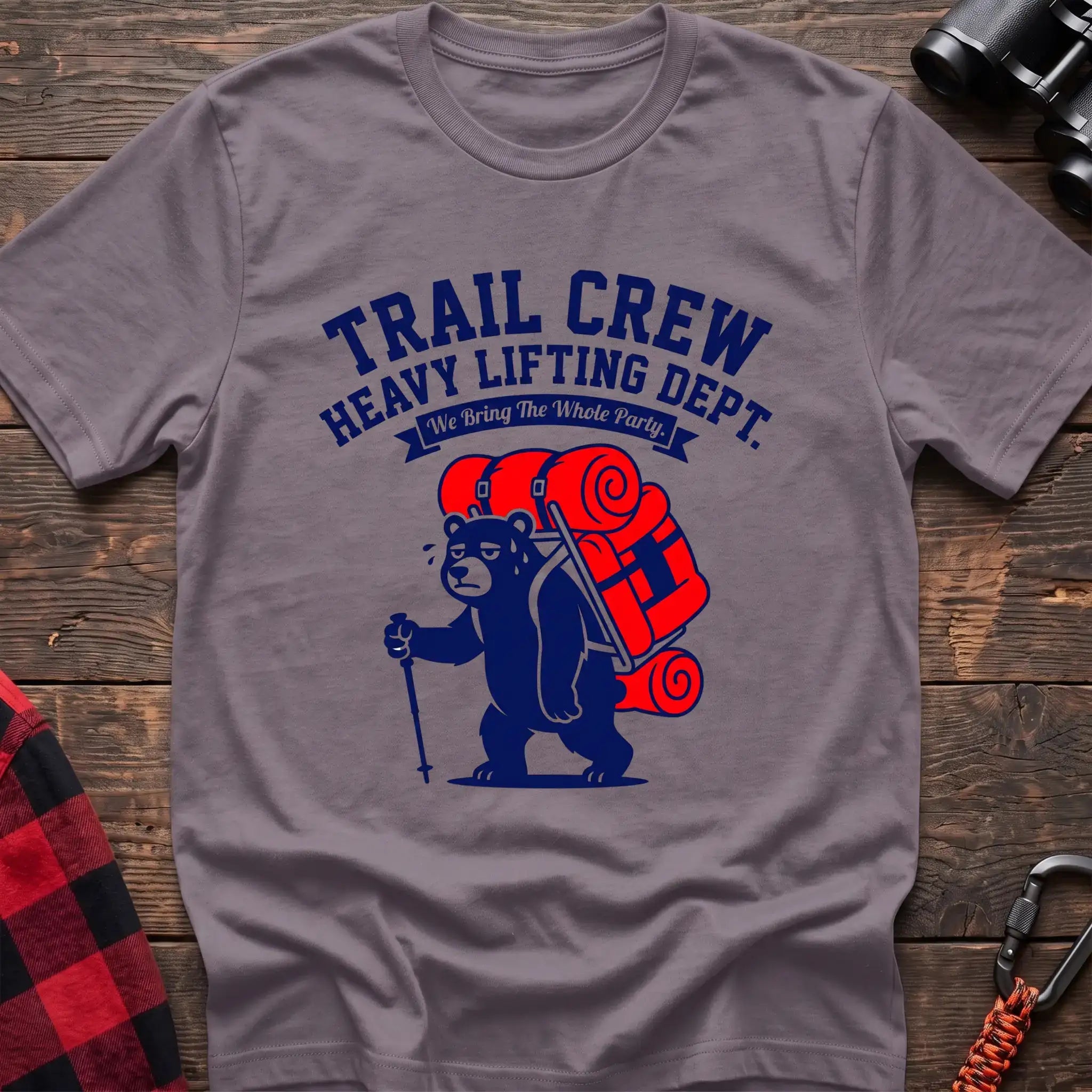 Paragon t-shirt with a cartoon dark blue bear carrying a large red backpack and hiking pole, with the text “TRAIL CREW HEAVY LIFTING DEPT. We Bring The Whole Party,” laid flat on a wooden surface.