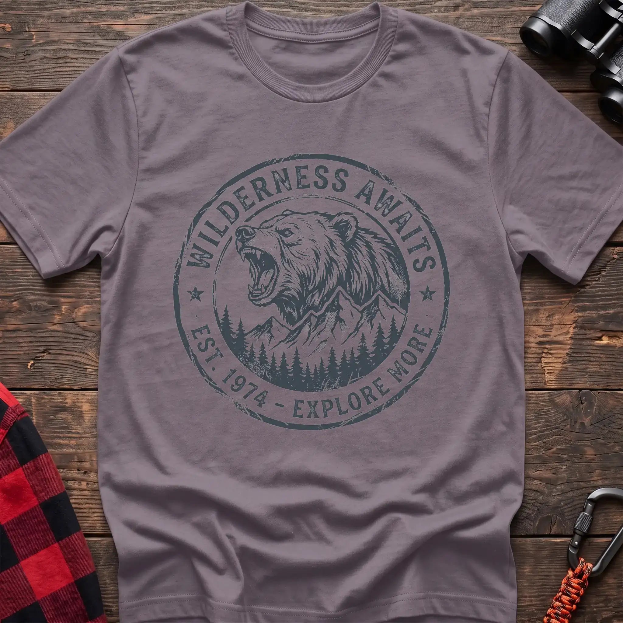 Paragon t-shirt with a circular dark gray badge design featuring a roaring bear head above mountains and pine trees, with the text “WILDERNESS AWAITS” and “EST. 1974 - EXPLORE MORE,” laid flat on a wooden surface.