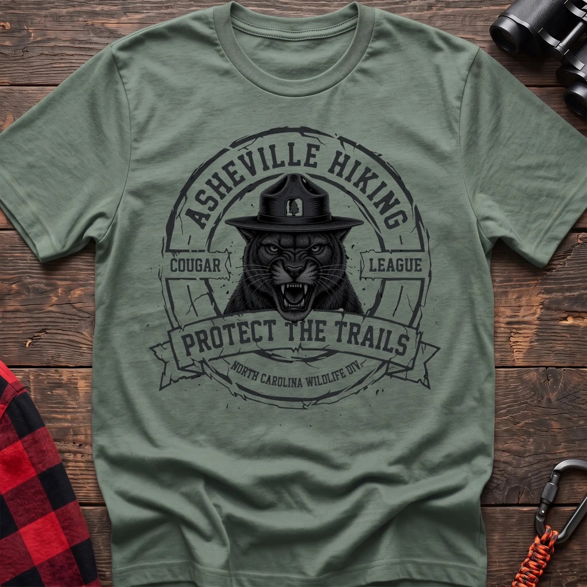 Sage cotton t-shirt with vintage Asheville Hiking Cougar League graphic of a roaring cougar in a park ranger hat, Protect the Trails North Carolina Wildlife Division badge, flat lay on rustic wood with hiking gear