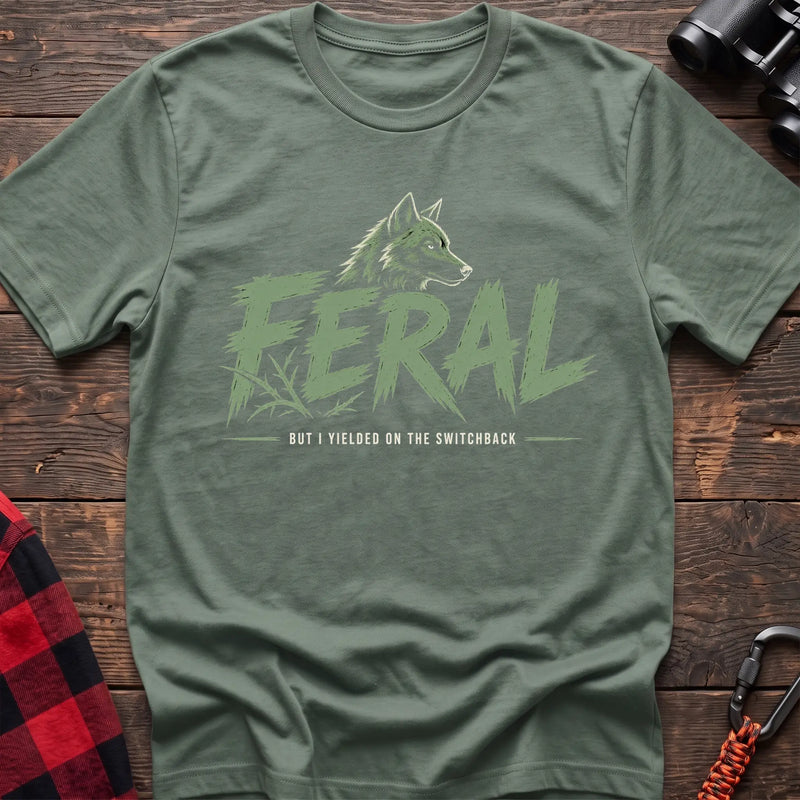 Sage cotton t-shirt with a wolf graphic and Feral But I Yielded on the Switchback text on rustic wood