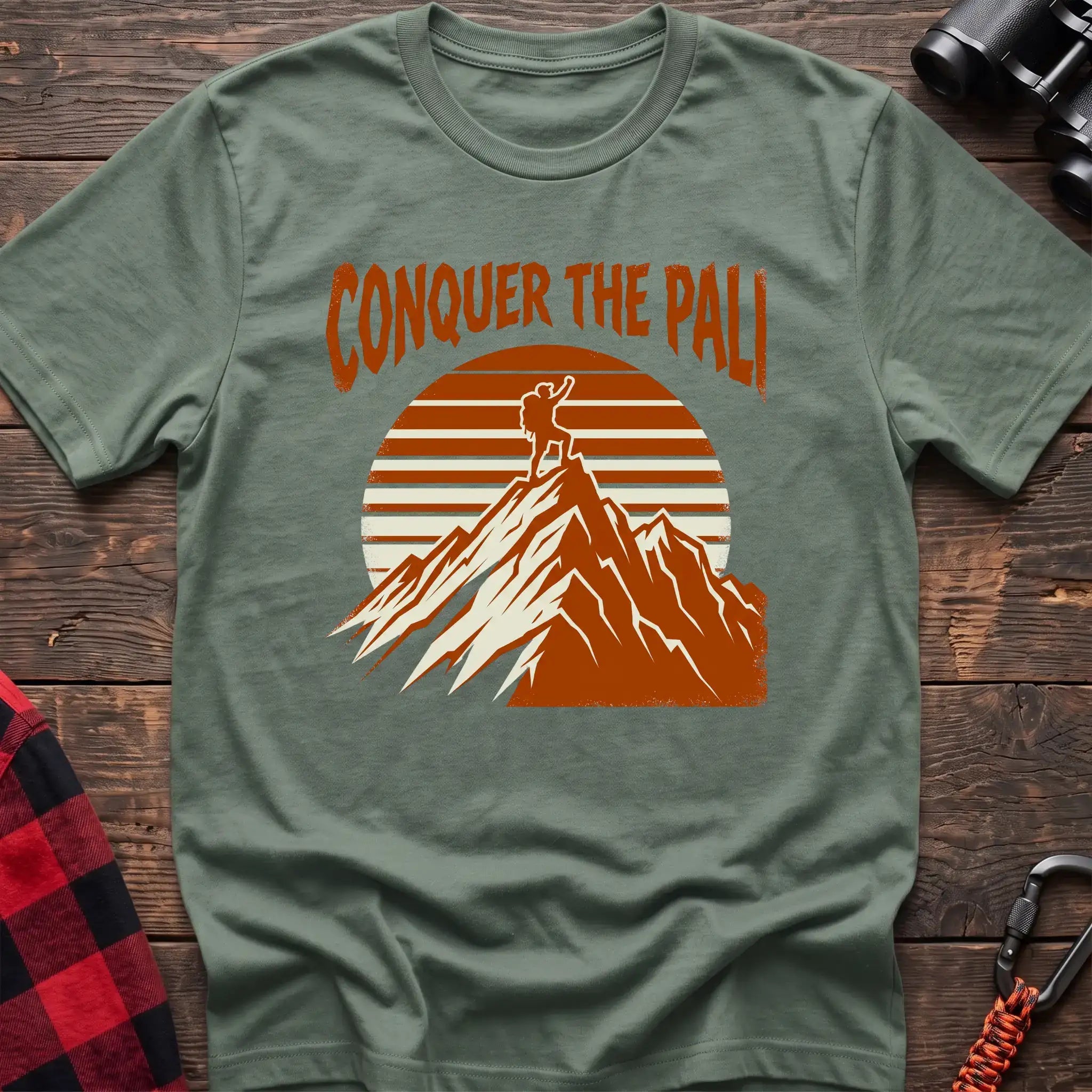 Sage t-shirt with orange text reading Conquer the Pali above a hiker standing on a mountain peak with a retro sun graphic