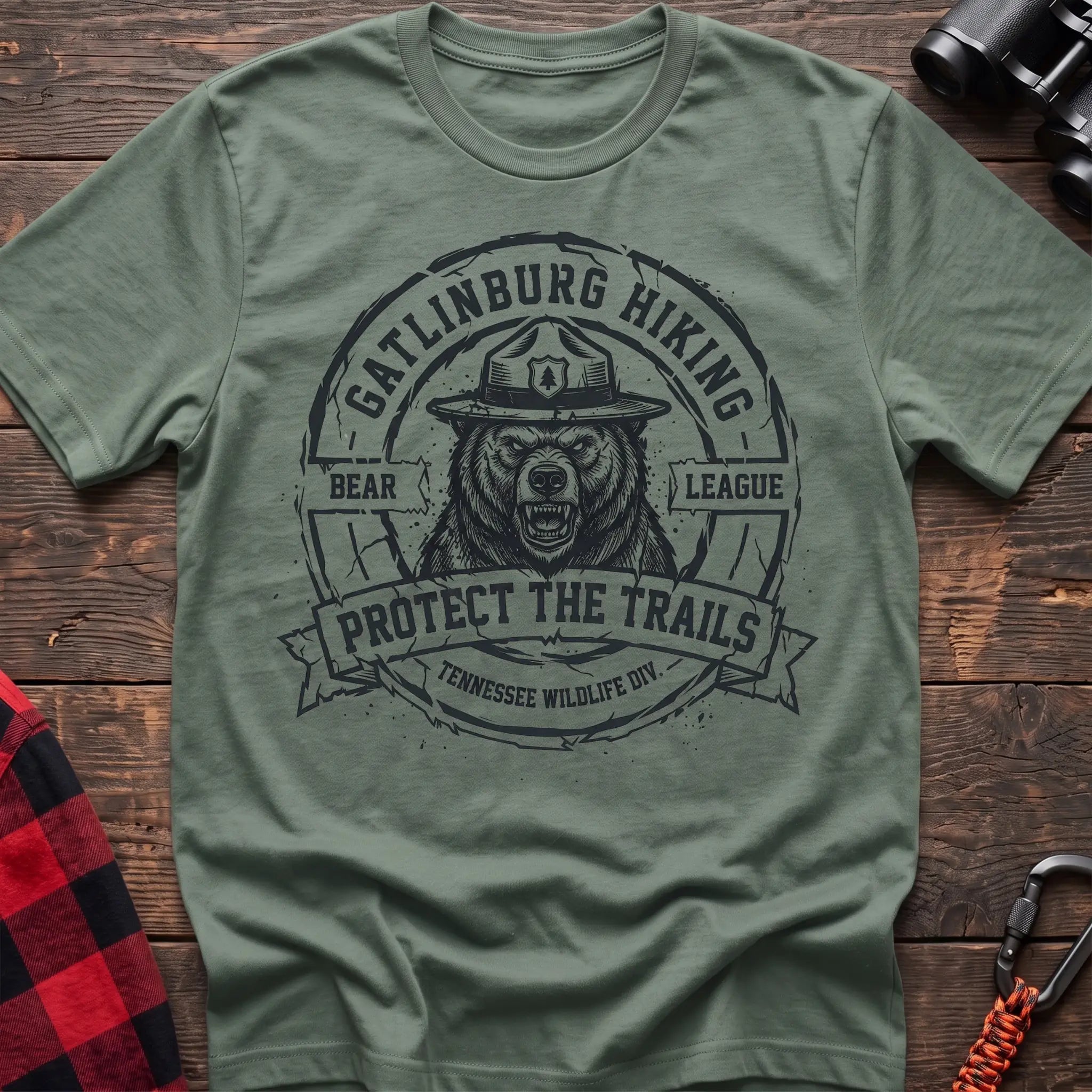 Sage cotton t-shirt with vintage Gatlinburg Hiking Bear League graphic of a roaring bear in a park ranger hat, Protect the Trails Tennessee Wildlife Division badge, flat lay on rustic wood with hiking gear