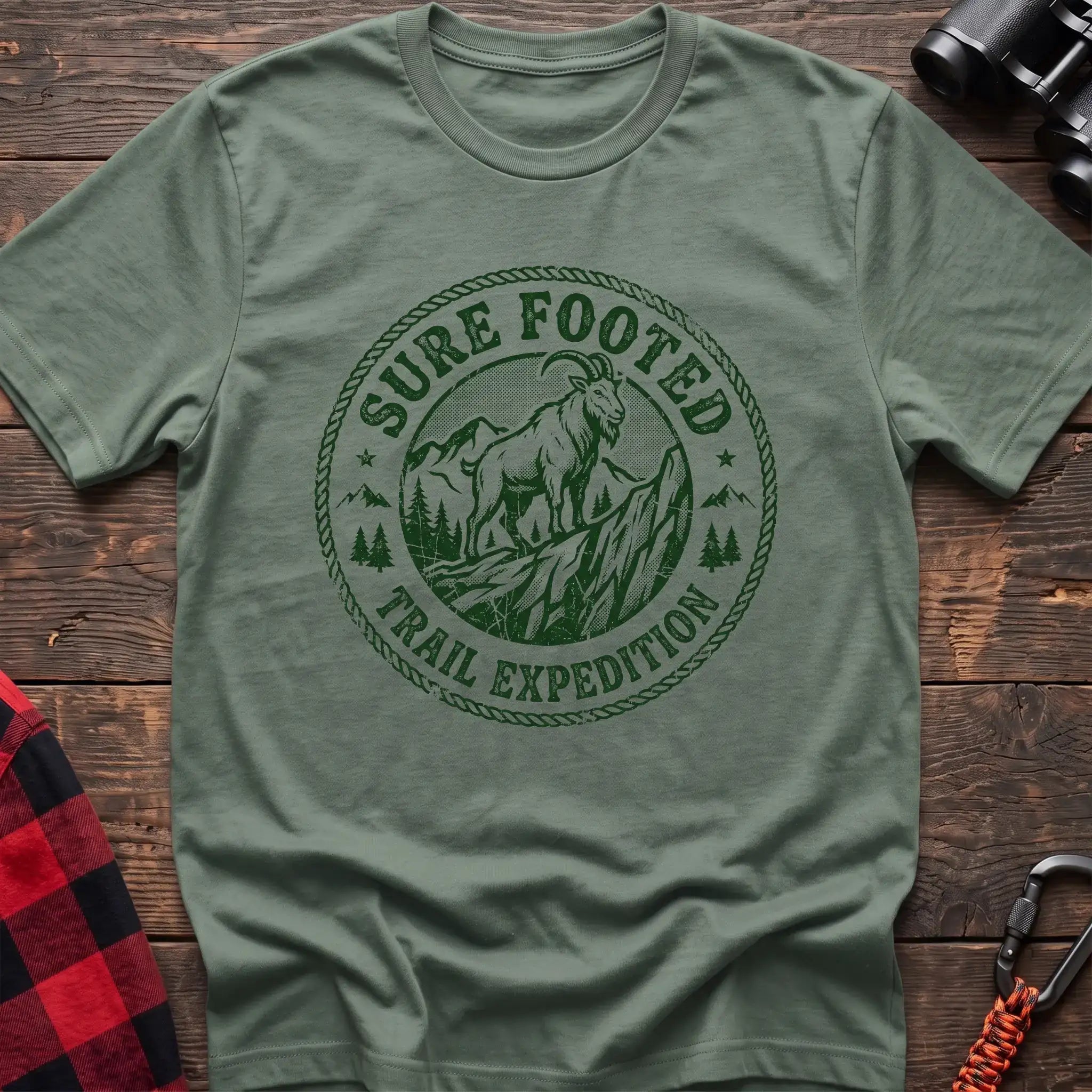 Sage green t-shirt with circular badge graphic of a mountain goat on a rocky peak reading Sure Footed Trail Expedition