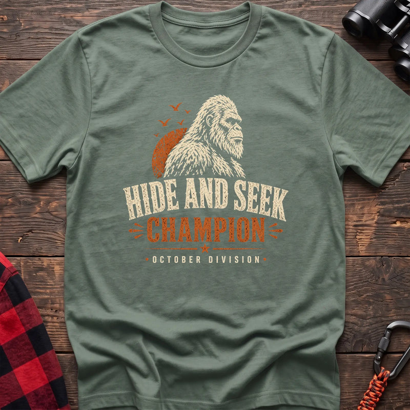 Sage cotton t-shirt with a Bigfoot graphic and Hide and Seek Champion October Division text on rustic wood