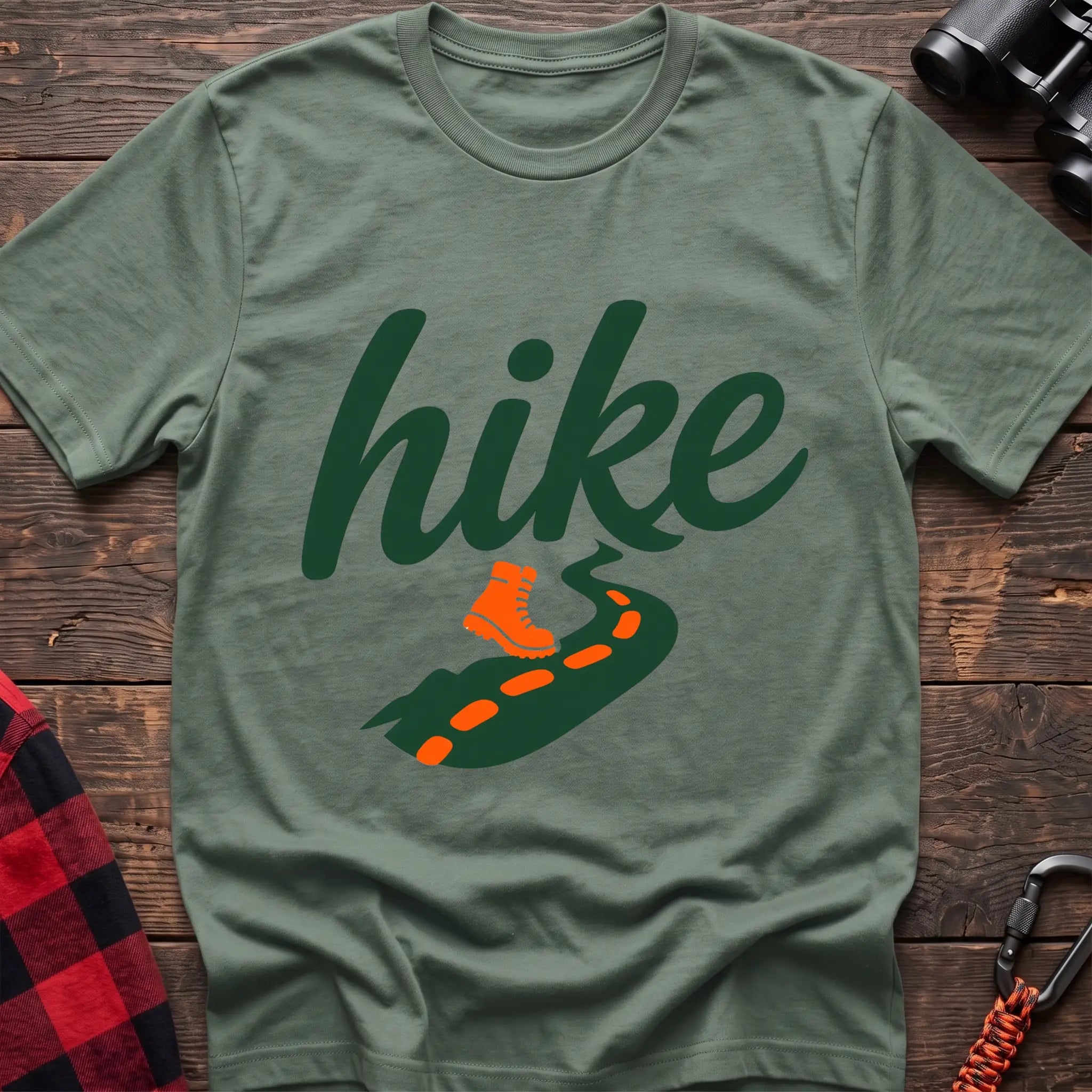 Sage cotton t-shirt with green cursive hike script, orange hiking boot, and footprint trail graphic, flat lay on rustic wood with hiking gear