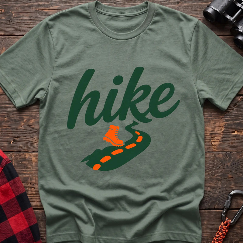 Sage cotton t-shirt with green cursive hike script, orange hiking boot, and footprint trail graphic, flat lay on rustic wood with hiking gear