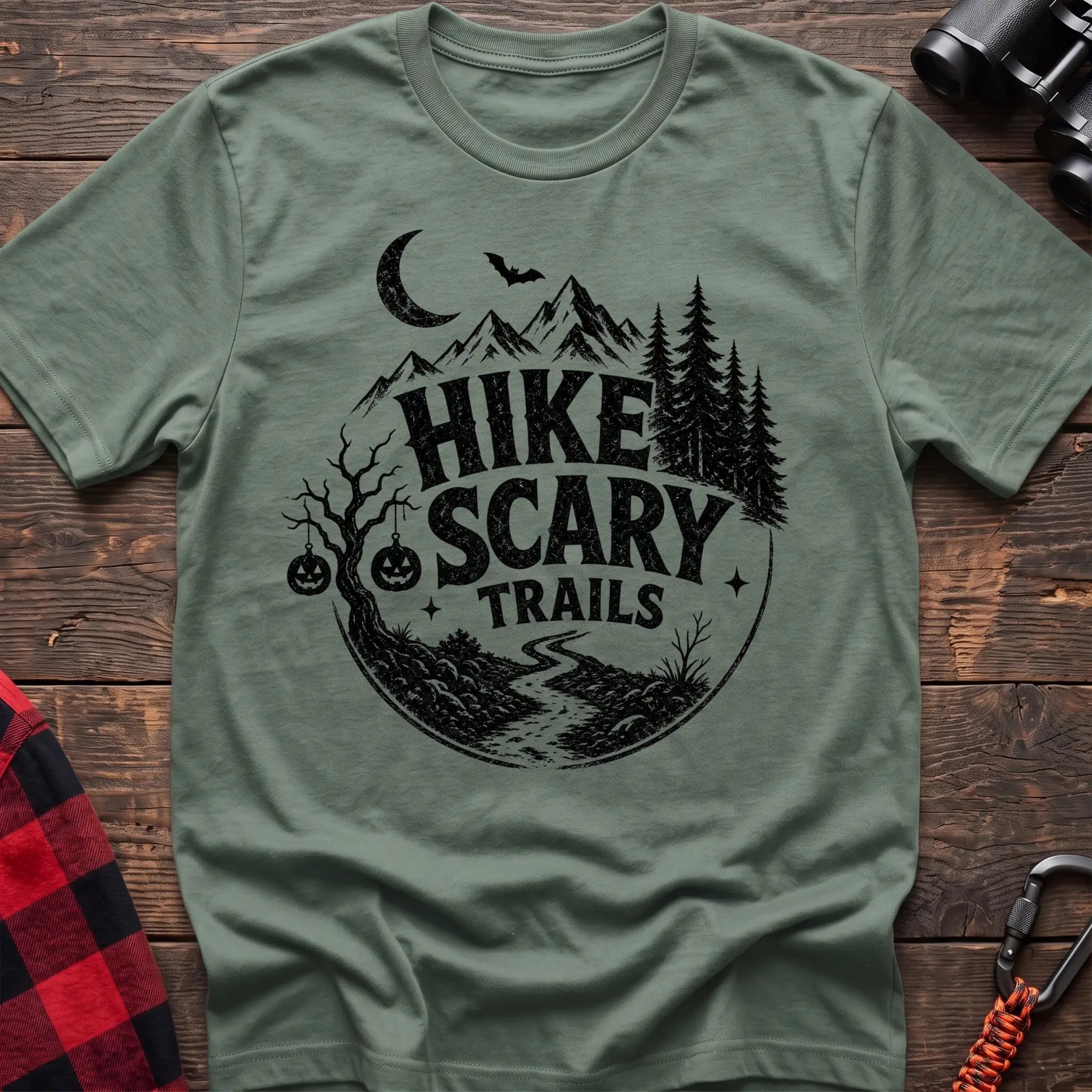 Sage green cotton t-shirt with vintage Hike Scary Trails graphic of mountains, a crescent moon, bat, jack-o-lanterns, and a winding trail, flat lay on rustic wood with hiking gear