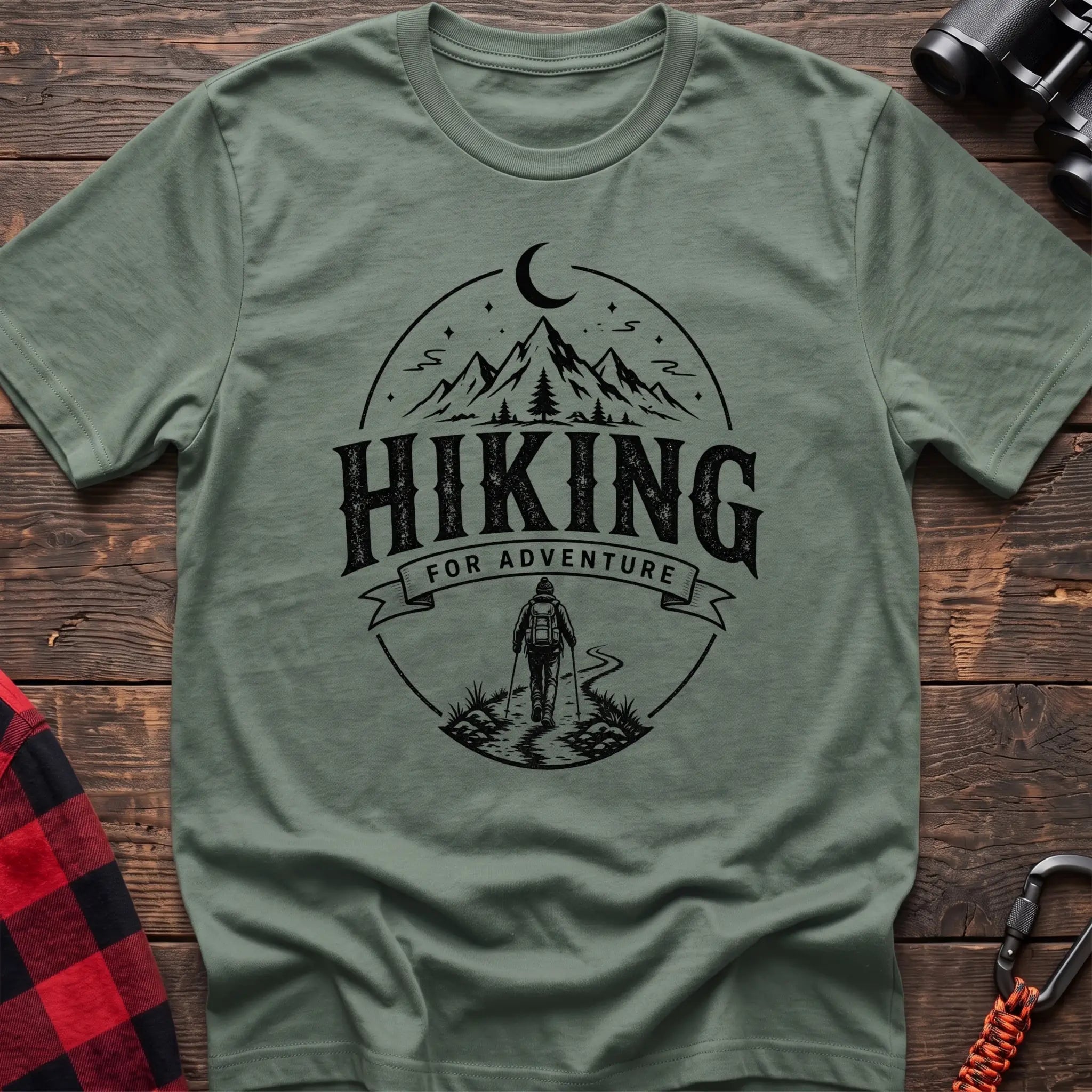 Sage green cotton t-shirt with vintage Hiking for Adventure graphic of mountains under a crescent moon and a hiker on a trail, flat lay on rustic wood with hiking gear