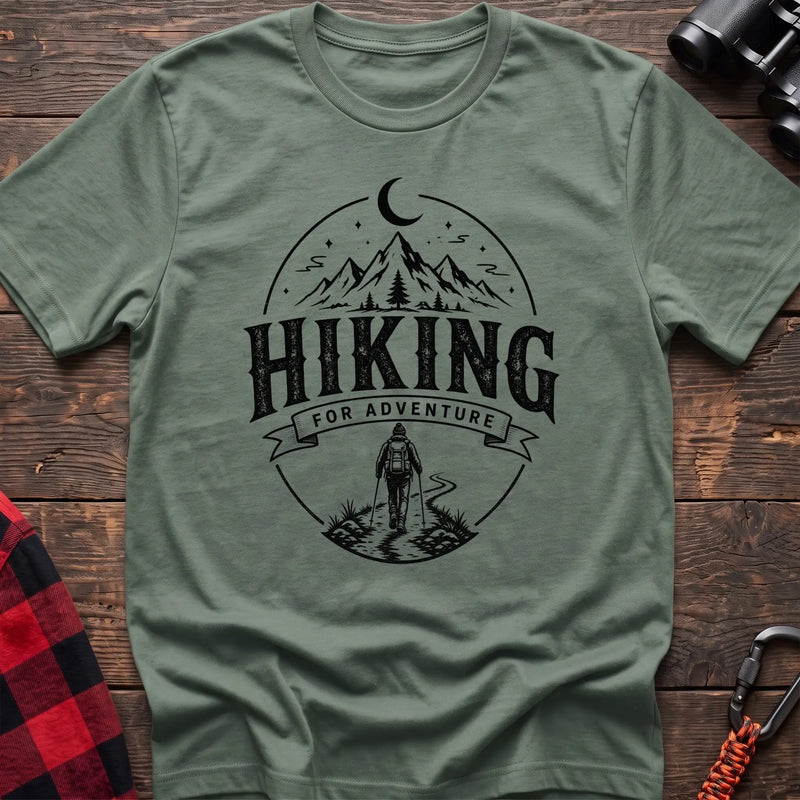 Sage green cotton t-shirt with vintage Hiking for Adventure graphic of mountains under a crescent moon and a hiker on a trail, flat lay on rustic wood with hiking gear