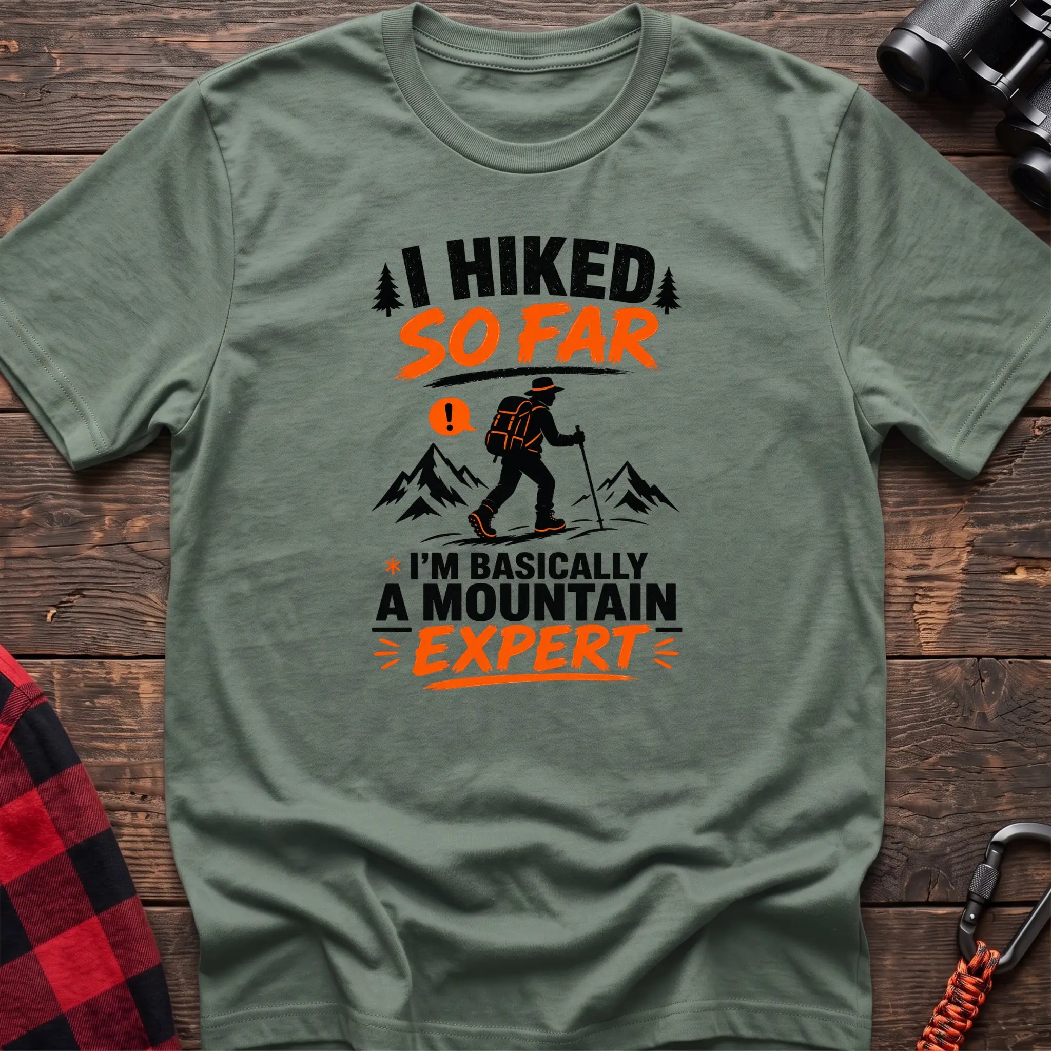 Sage cotton t-shirt with I Hiked So Far I'm Basically a Mountain Expert graphic of a hiker with backpack and trekking pole, flat lay on rustic wood with hiking gear