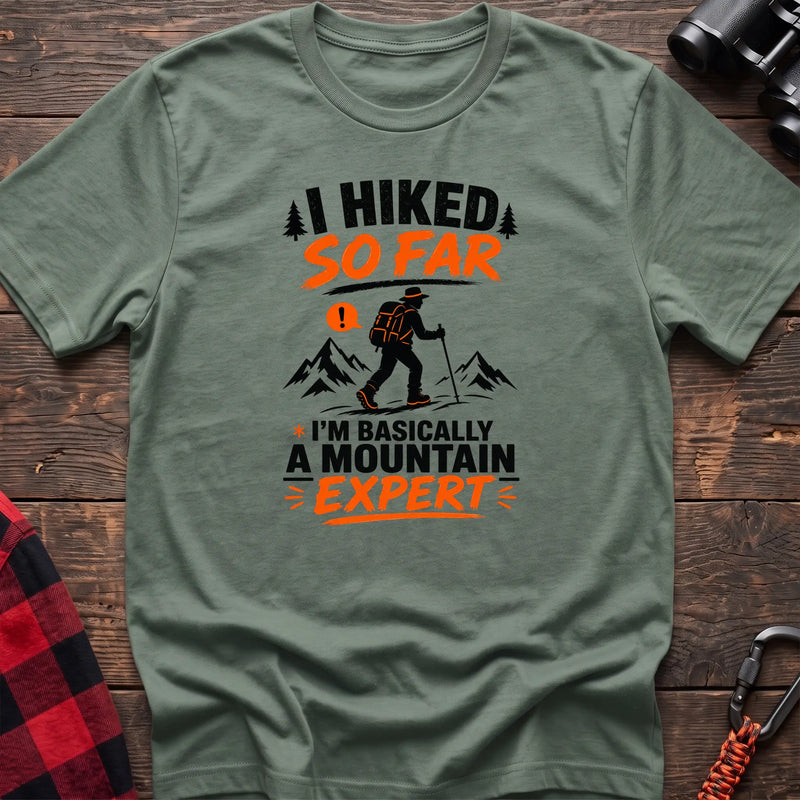 Sage cotton t-shirt with I Hiked So Far I'm Basically a Mountain Expert graphic of a hiker with backpack and trekking pole, flat lay on rustic wood with hiking gear