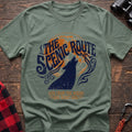 Sage green t-shirt with a dark blue howling wolf silhouette against an orange circle, with the arched text “THE SCENIC ROUTE” above and “LIVE FROM THE WOODS / ONE NIGHT ONLY” below, laid flat on a wooden surface.