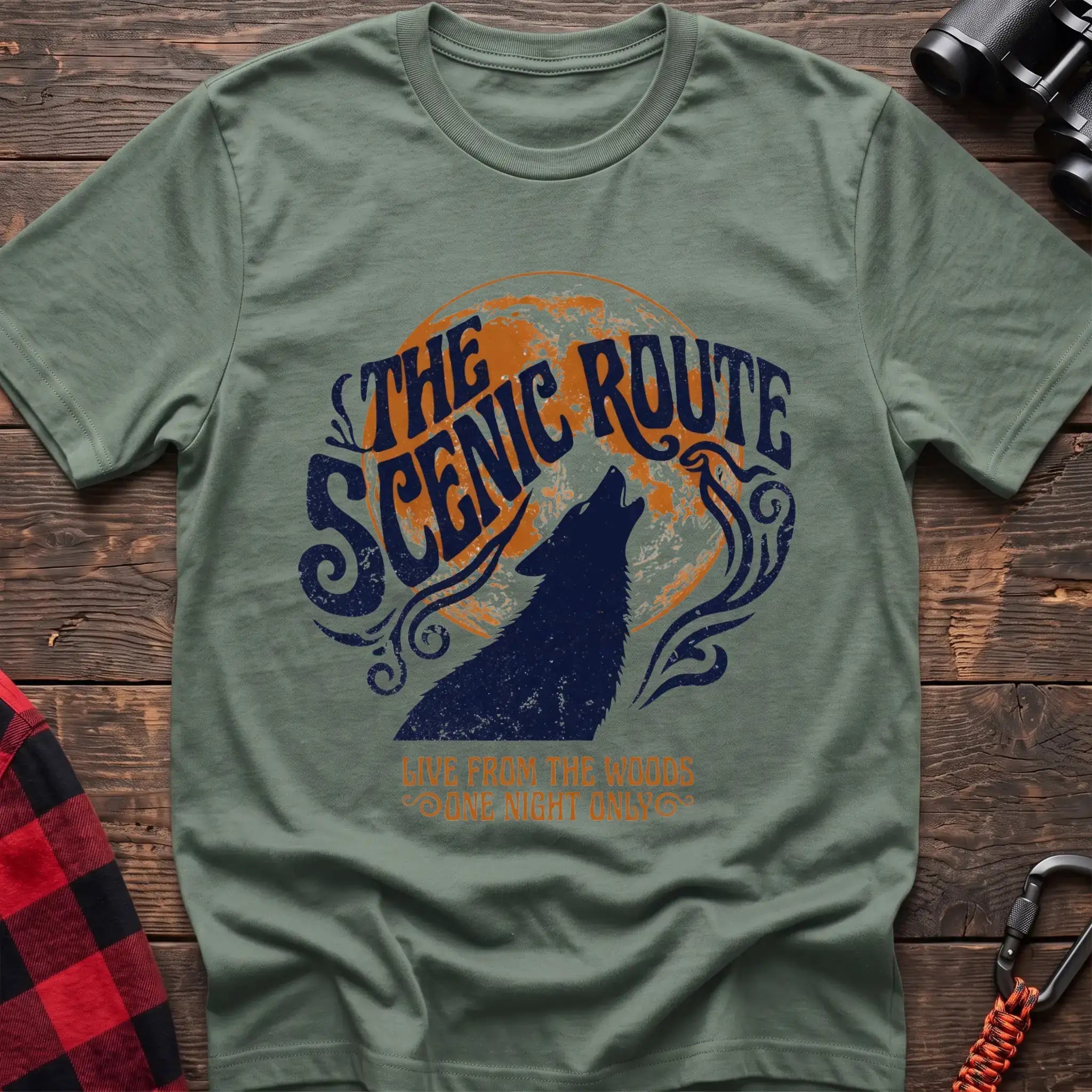 Sage green t-shirt with a dark blue howling wolf silhouette against an orange circle, with the arched text “THE SCENIC ROUTE” above and “LIVE FROM THE WOODS / ONE NIGHT ONLY” below, laid flat on a wooden surface.