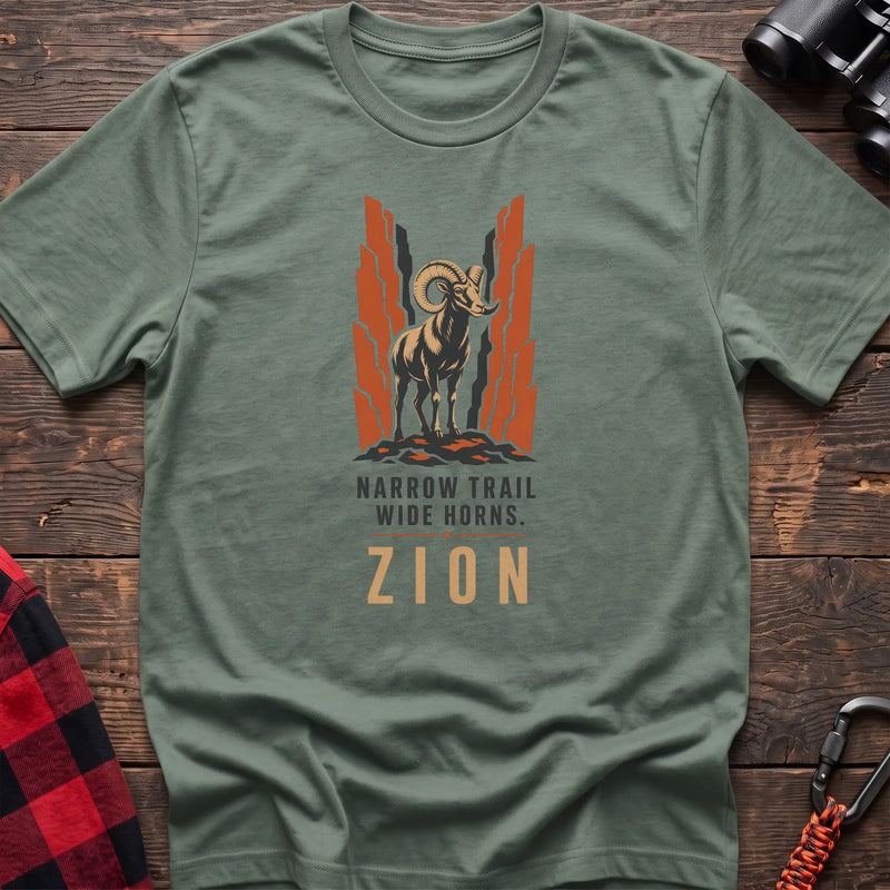 Sage cotton t-shirt with a bighorn sheep graphic and Narrow Trail Wide Horns Zion text on rustic wood